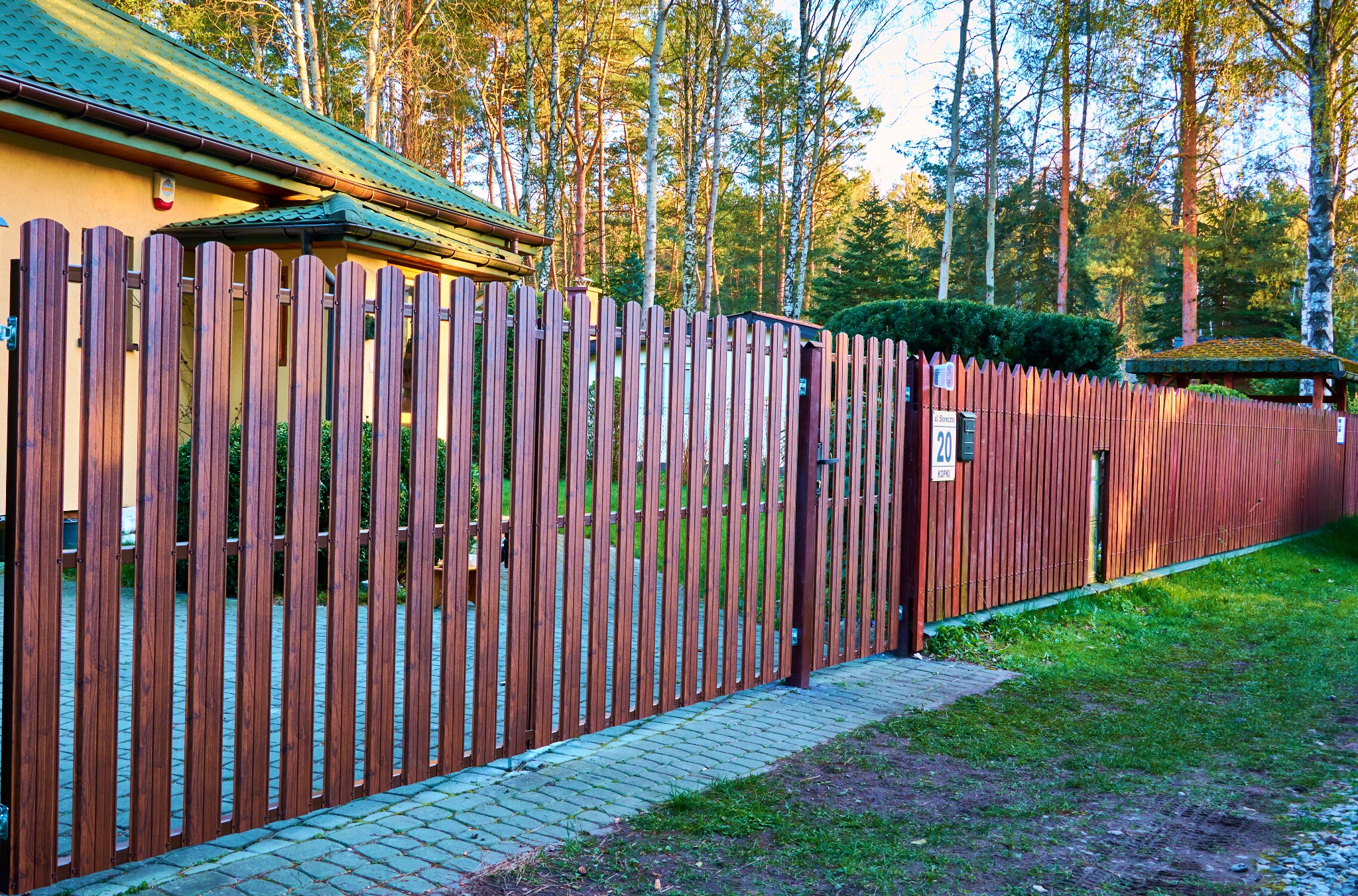 Brązowe ogrodzenie panelowe z furtką, widok z zewnątrz posesji, z numerem domu na słupku.