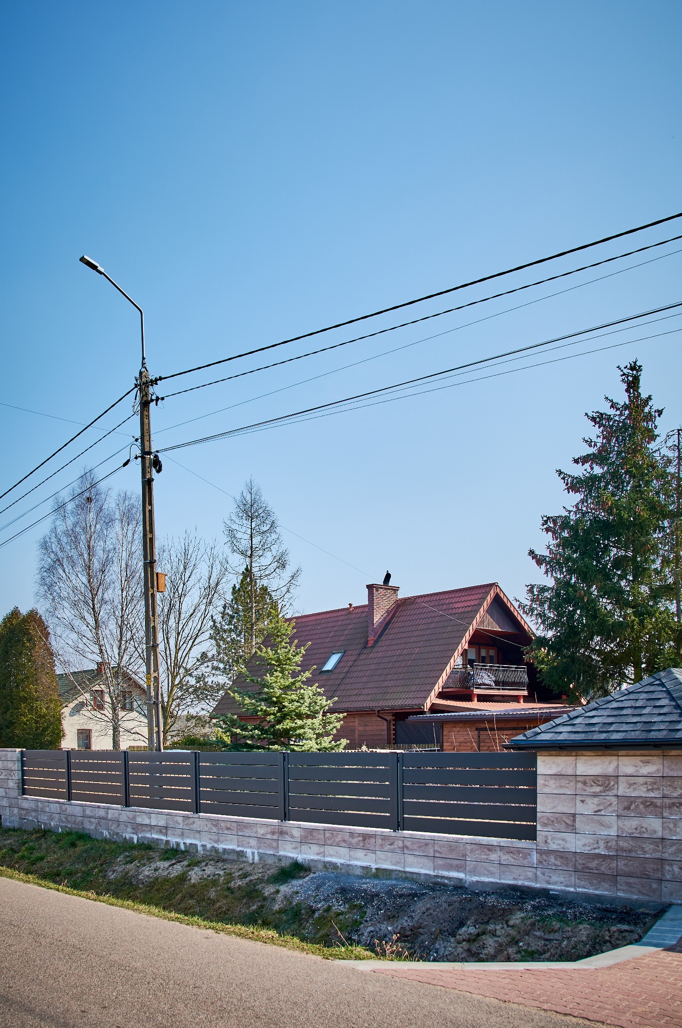Nowoczesne, poziome przęsła ogrodzeniowe w kolorze grafitowym, osadzone na murku z beżowych bloczków imitujących kamień, na tle domu z czerwoną dachówką i zielonymi drzewami w słoneczny dzień.