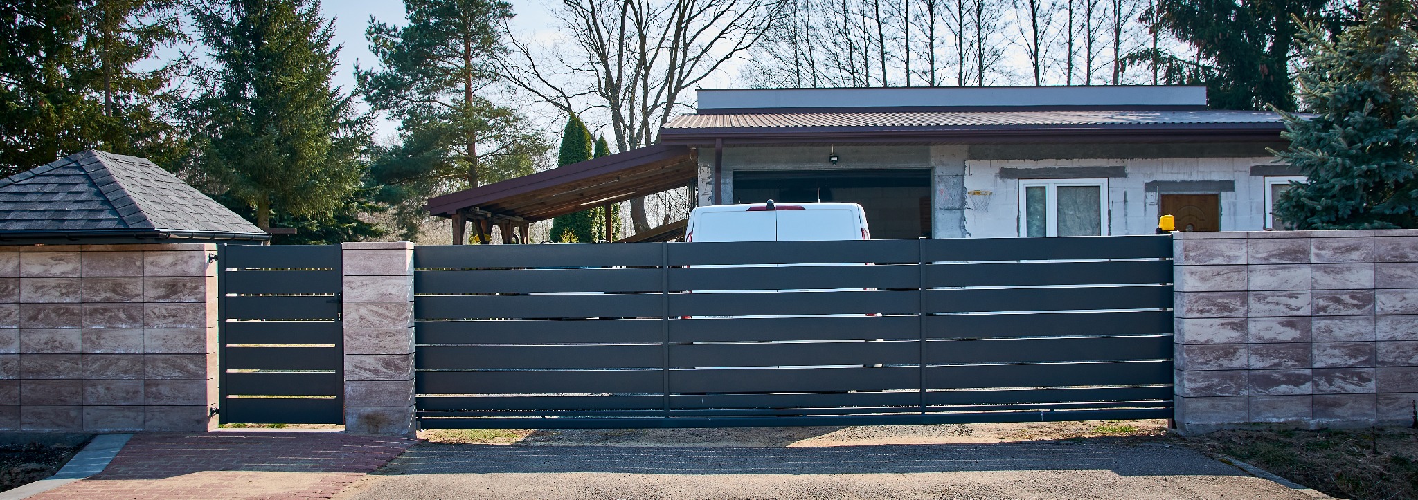 Nowoczesna, przesuwna brama z poziomych, szarych profili, osadzona na słupkach z dekoracyjnych, brązowo-beżowych bloczków betonowych. W tle widoczny budynek mieszkalny z białym vanem zaparkowanym...