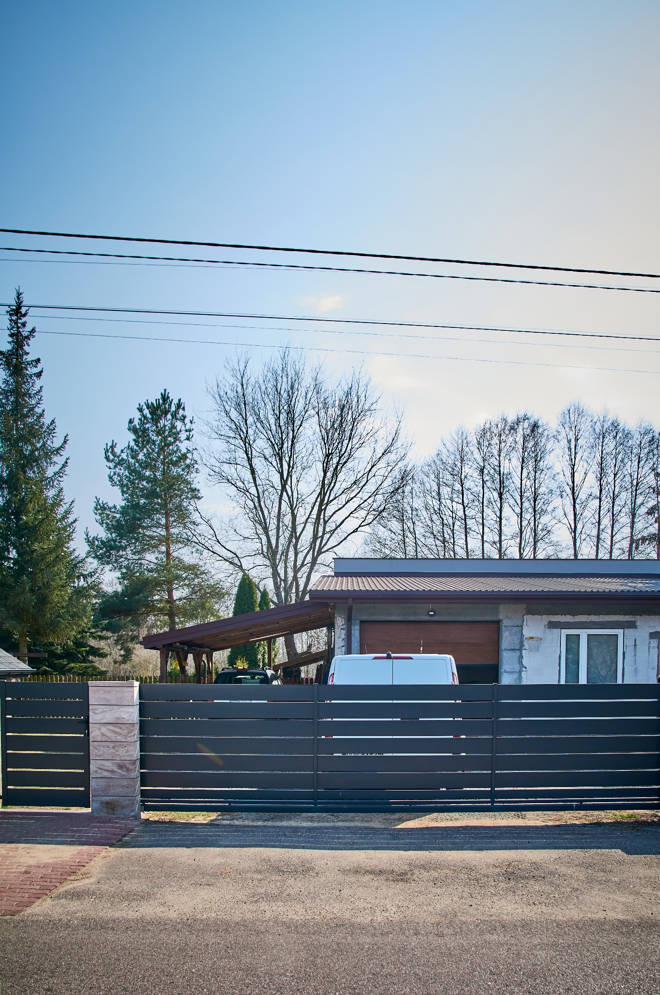 Nowoczesna, pozioma brama wjazdowa z szarych paneli metalowych, z jasnymi słupkami z imitacji kamienia, na tle domu z garażem i podjazdem, w słoneczny dzień.