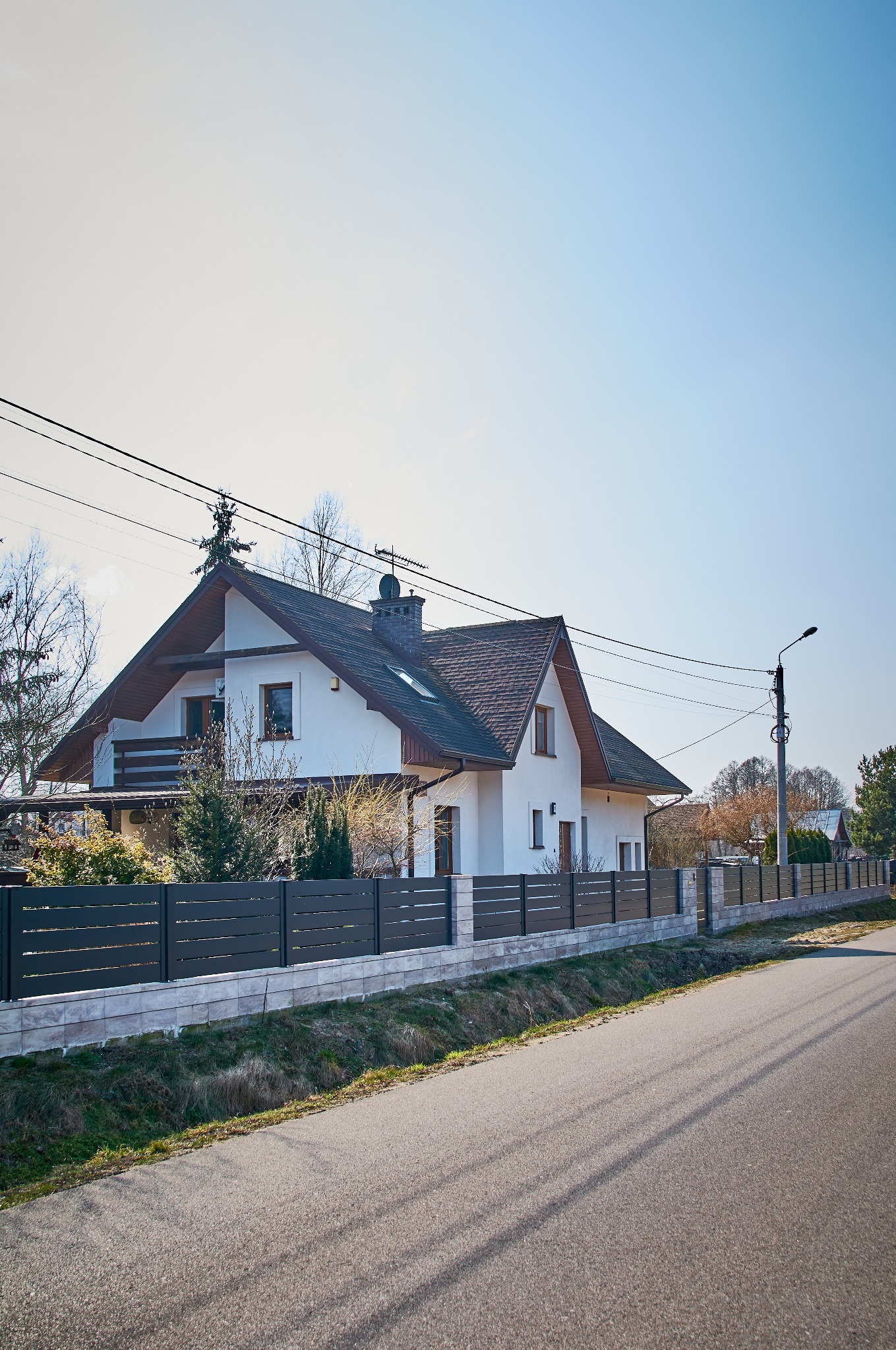 Nowoczesny dom jednorodzinny z ciemnoszarym, poziomym ogrodzeniem panelowym na podmurówce z jasnych płytek, widziany z perspektywy ulicy w słoneczny dzień.