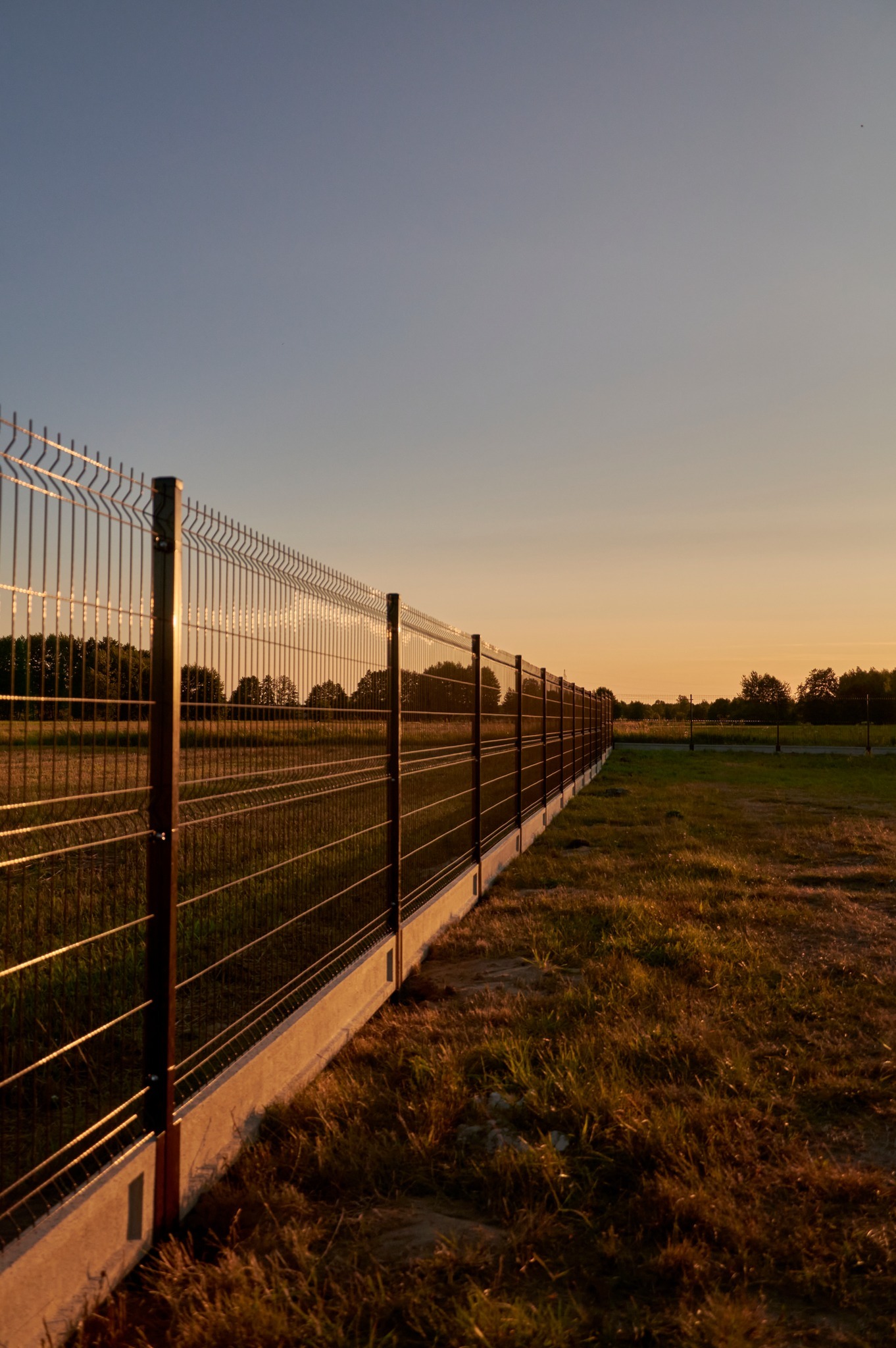 Długi, brązowy płot panelowy z podmurówką betonową oddzielający pole od łąki, sfotografowany pod kątem podczas zachodu słońca.