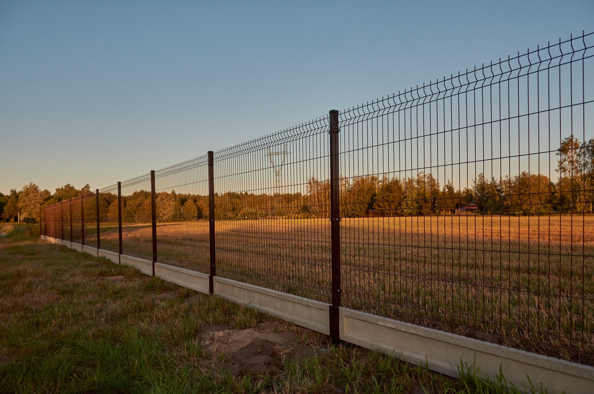 Długi, ciemny, metalowy płot panelowy z podmurówką betonową, rozciągający się wzdłuż pola w promieniach zachodzącego słońca.
