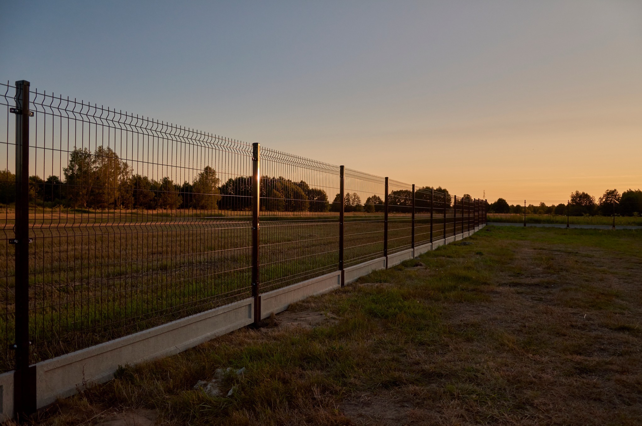 Długi, ciemny, metalowy płot panelowy z betonową podmurówką, rozciągający się wzdłuż pola o zmierzchu, z drzewami w tle.