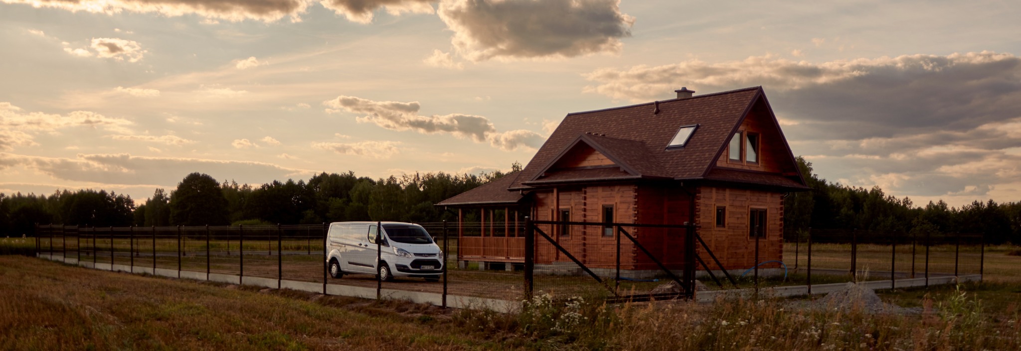 Nowoczesny dom z bali otoczony panelowym ogrodzeniem, z białym vanem zaparkowanym na podjeździe, w tle las i zachmurzone niebo.