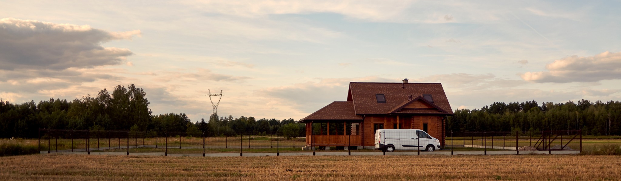 Nowoczesny dom z drewna otoczony niskim, ciemnym ogrodzeniem panelowym na tle pola i lasu, z zaparkowanym białym vanem dostawczym.