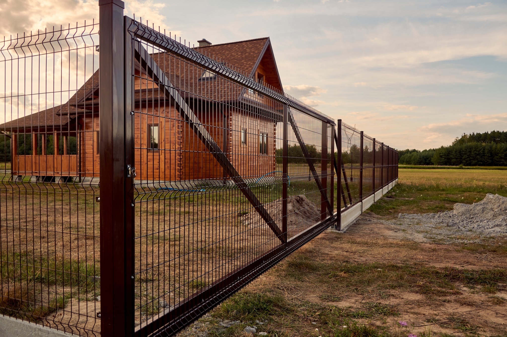 Ciemnobrązowe ogrodzenie panelowe z bramą wjazdową, widok na drewniany dom z brązowym dachem w tle, słoneczny dzień.