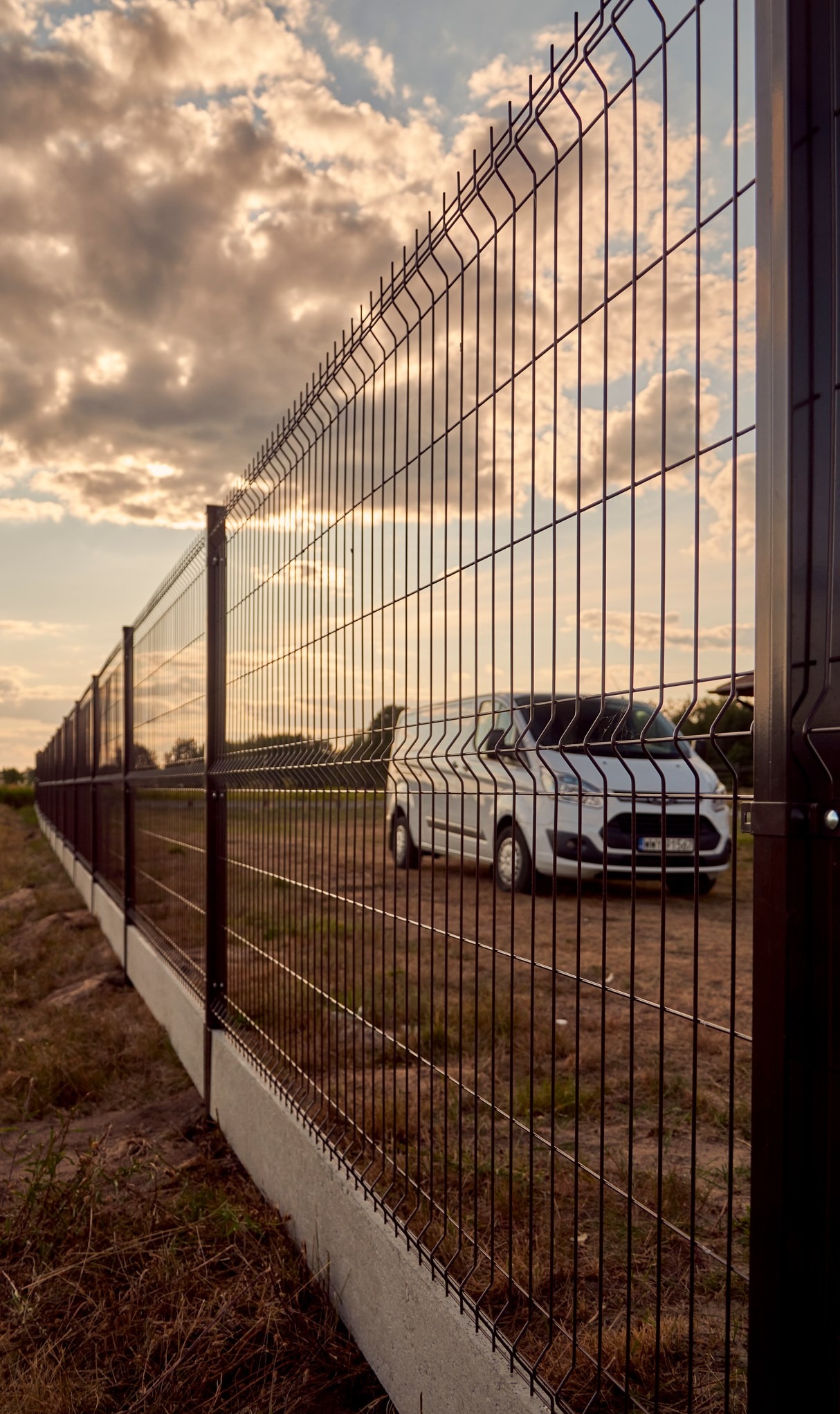 Ciemnobrązowe ogrodzenie panelowe na betonowej podmurówce, z białym dostawczym samochodem widocznym w tle i zachodzącym słońcem tworzącym ciepłą poświatę na chmurach.