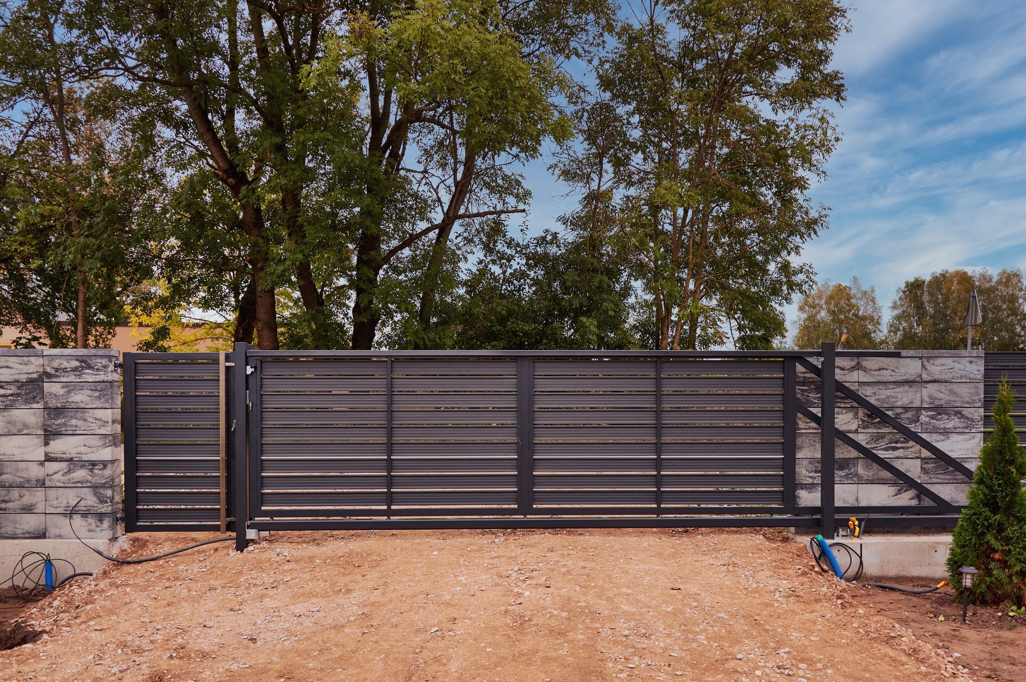 Nowoczesna brama wjazdowa z poziomymi panelami, zamontowana między betonowymi słupkami, z widocznymi przewodami i elementami instalacji elektrycznej na gruncie.