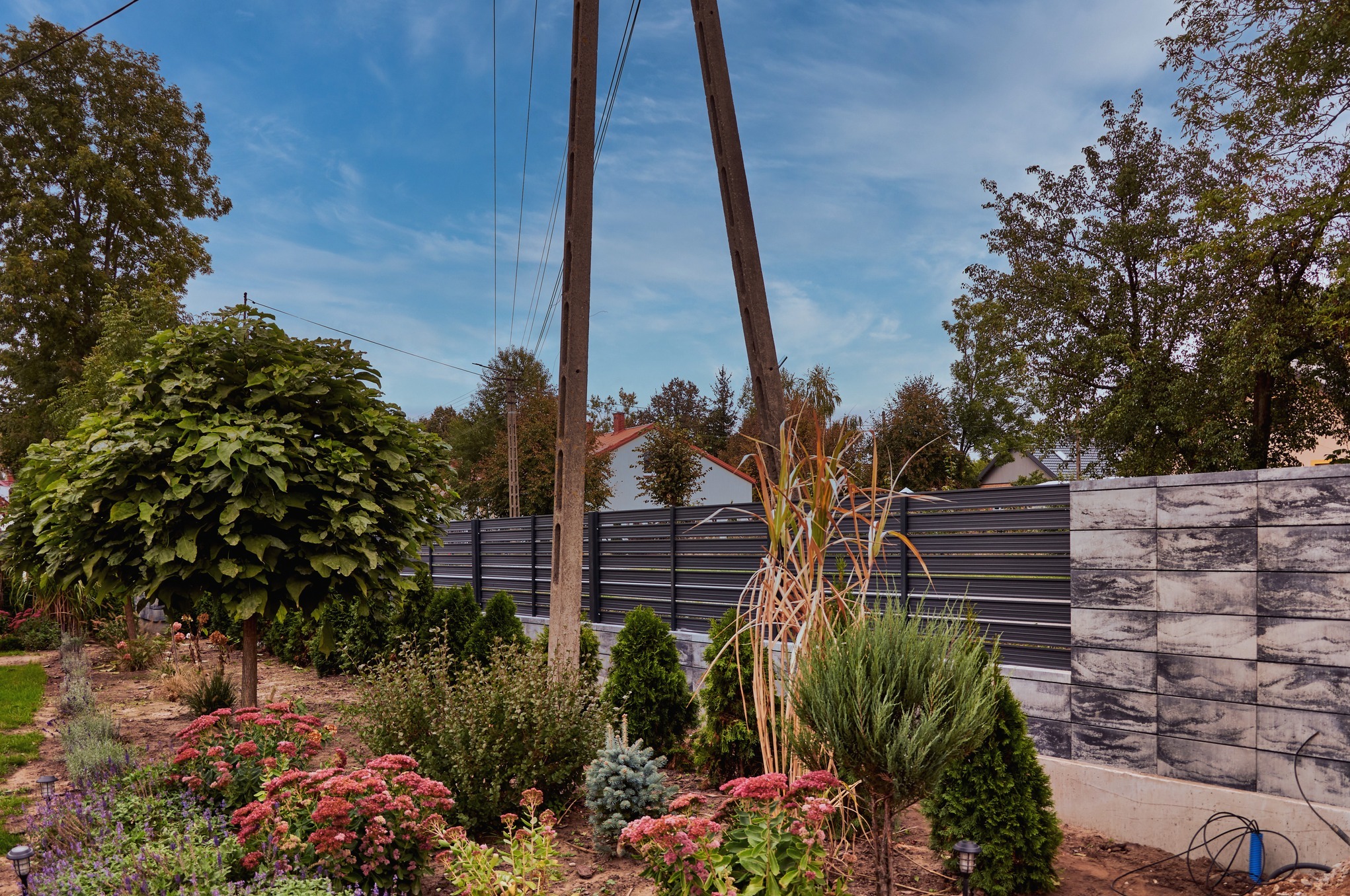 Nowoczesne ogrodzenie panelowe w kolorze antracytowym z podmurówką imitującą kamień, widoczne na tle zadbanego ogrodu z kwitnącymi roślinami i drzewami.