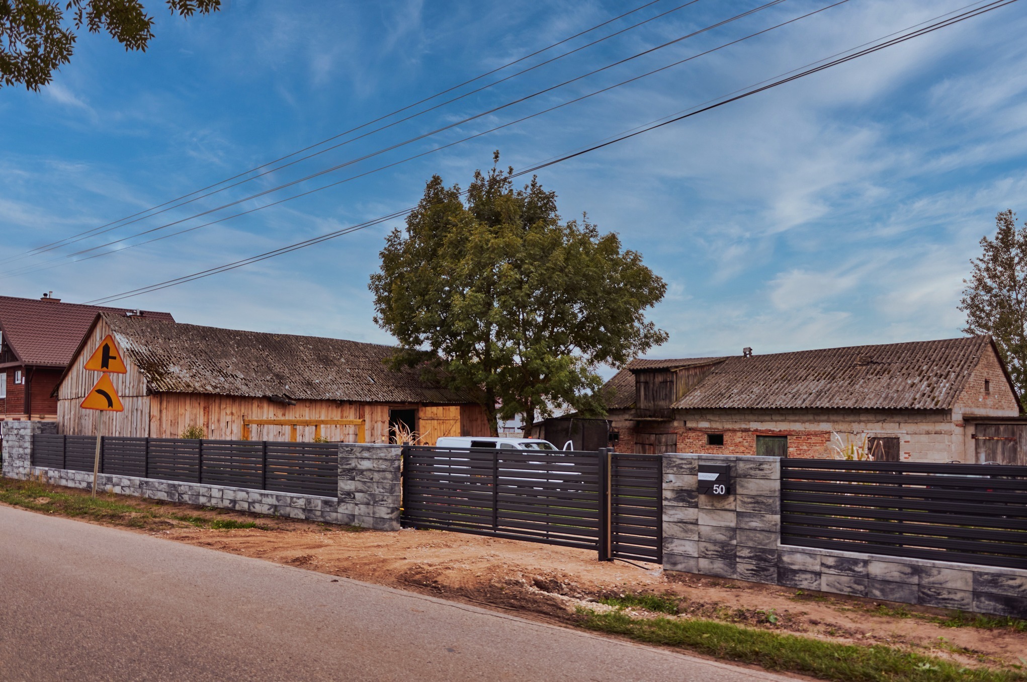 Nowoczesna, pozioma brama wjazdowa i przęsła ogrodzeniowe z szarego metalu z numerem 50 na słupku, z betonową podmurówką w kolorze grafitu, na tle wiejskich zabudowań i błękitnego nieba.