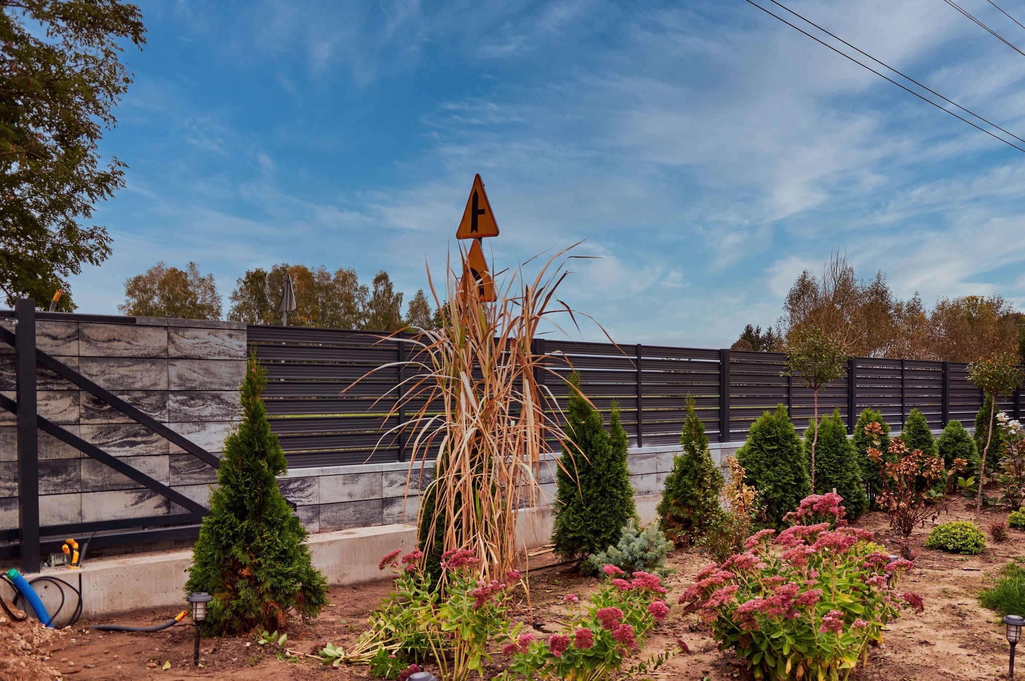 Nowoczesne ogrodzenie panelowe z elementami betonowymi, widok na ogród z tujami i kwiatami, w tle znak drogowy.