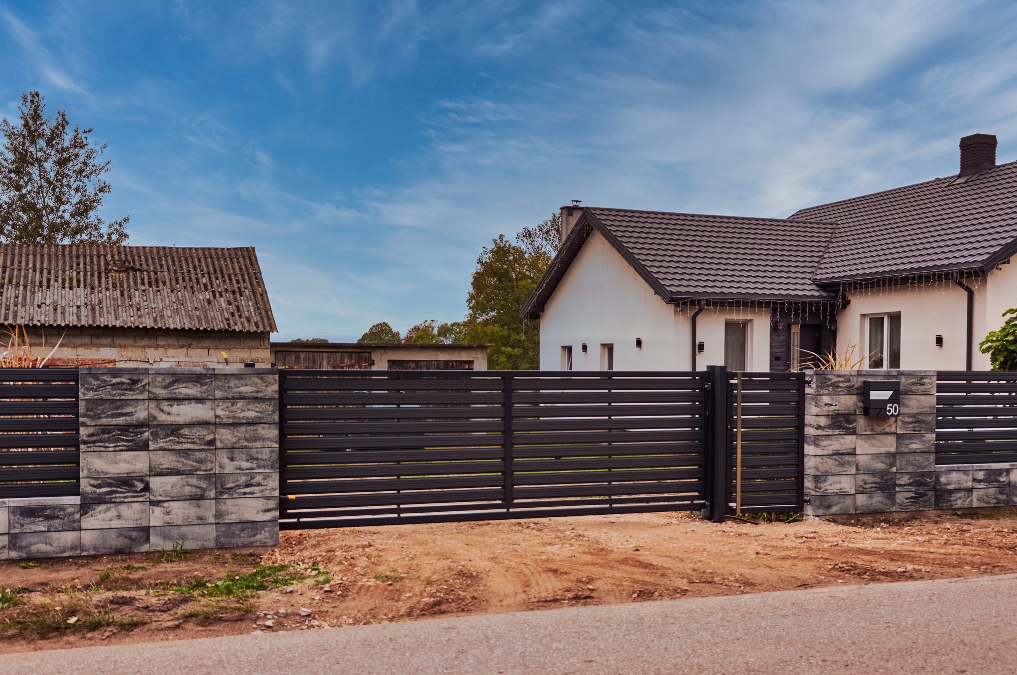 Nowoczesne, poziome przęsła w kolorze antracytowym tworzą ogrodzenie posesji z jasnym domem i starą stodołą w tle. Słupki ogrodzeniowe z szarej kostki betonowej. Skrzynka pocztowa z numerem 50.