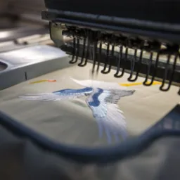 Automatyczna maszyna do haftowania w trakcie pracy, igła precyzyjnie tworzy wzór lecącego żurawia na beżowym materiale, widoczne detale mechanizmu i napięta nić.