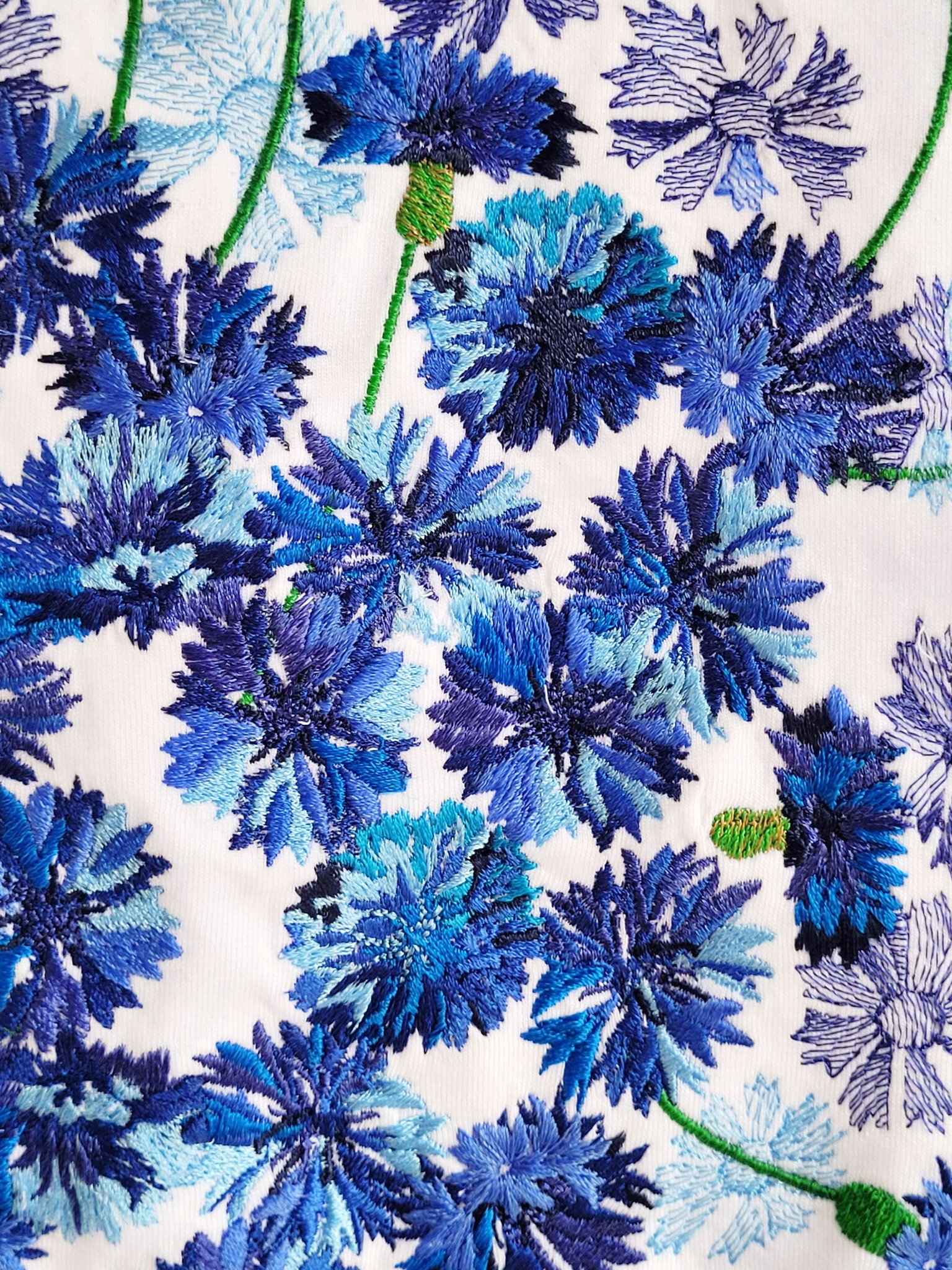 Delikatny haft przedstawiający niebieskie chabry na białym materiale, zróżnicowane odcienie nici tworzą trójwymiarowy efekt.