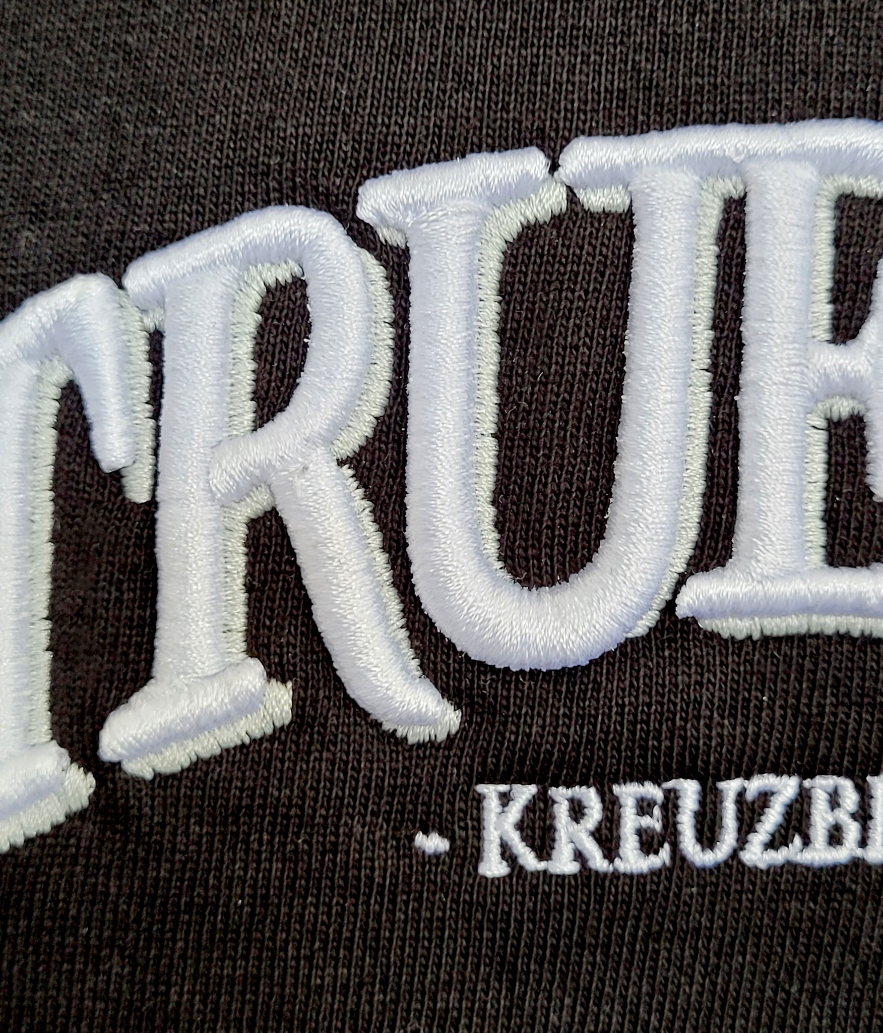 Zbliżenie na fragment ciemnobrązowej tkaniny z białym, trójwymiarowym haftem napisu 'TRUE - KREUZBI'