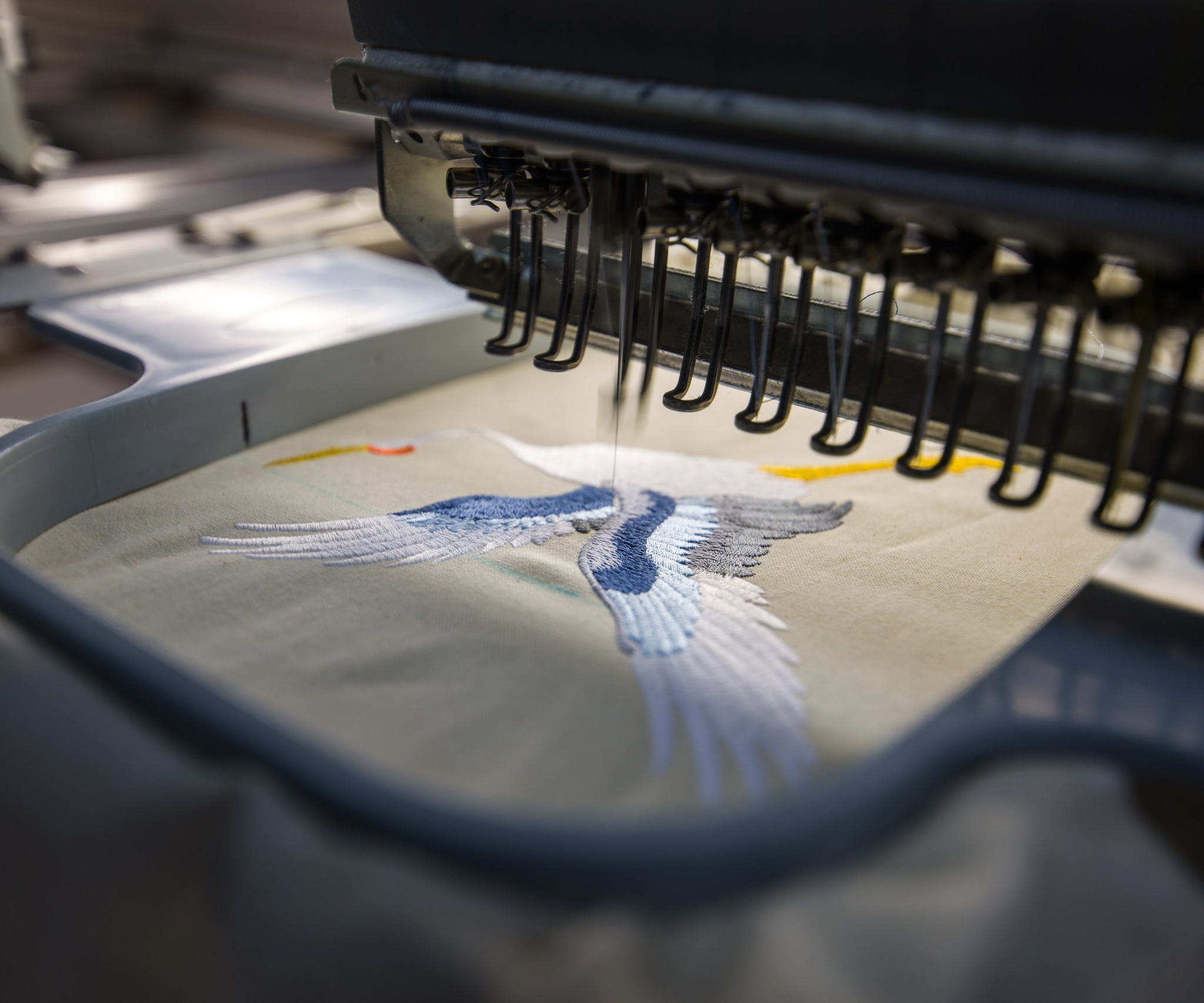 Automatyczna maszyna do haftowania w trakcie pracy, igła precyzyjnie tworzy wzór lecącego żurawia na beżowym materiale, widoczne detale mechanizmu i napięta nić.