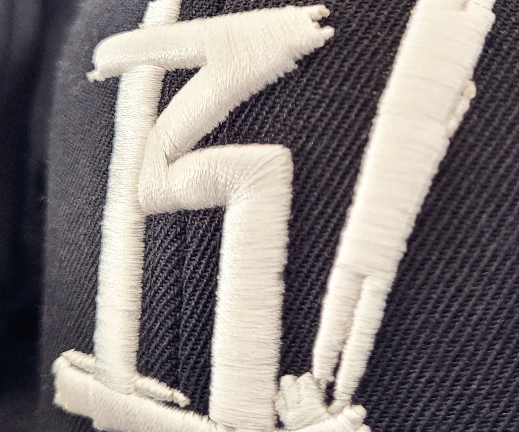 Detal haftu 3D na ciemnej tkaninie, przedstawiający białe, wypukłe litery z bliska, ukazujące strukturę nici i materiału.