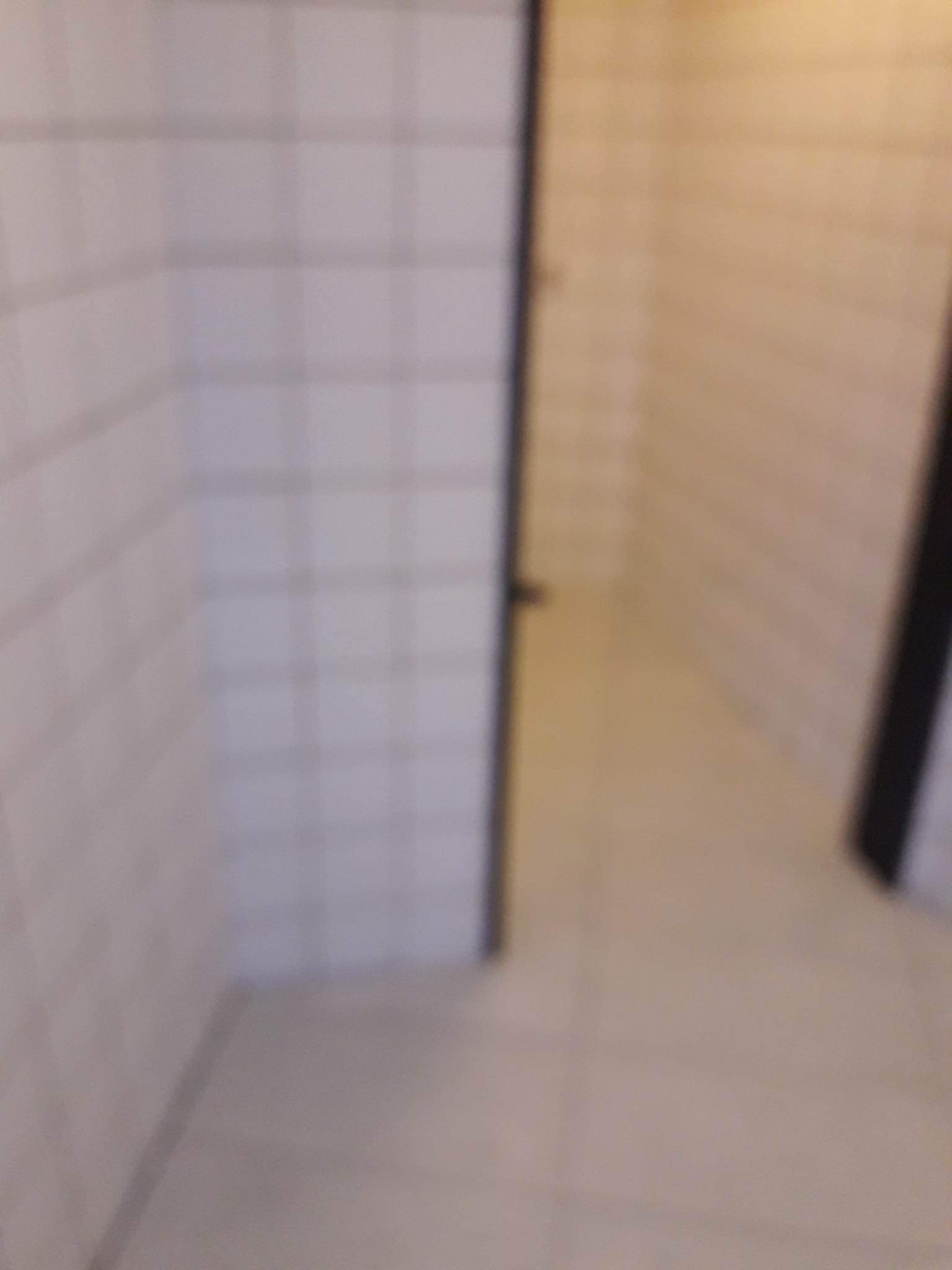 Rozmyte ujęcie fragmentu łazienki z białymi kwadratowymi płytkami na ścianach i jasną podłogą, widoczny fragment czarnej ramy drzwi.