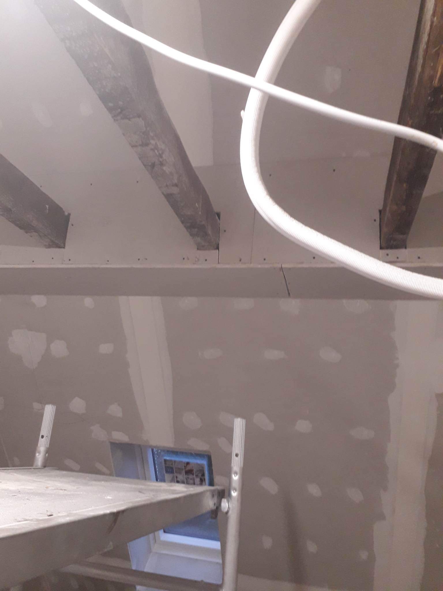 Wnętrze w trakcie prac wykończeniowych: surowa ściana z widocznymi łatami gipsowymi, drewniane belki stropowe, biała rura instalacyjna i fragment rusztowania