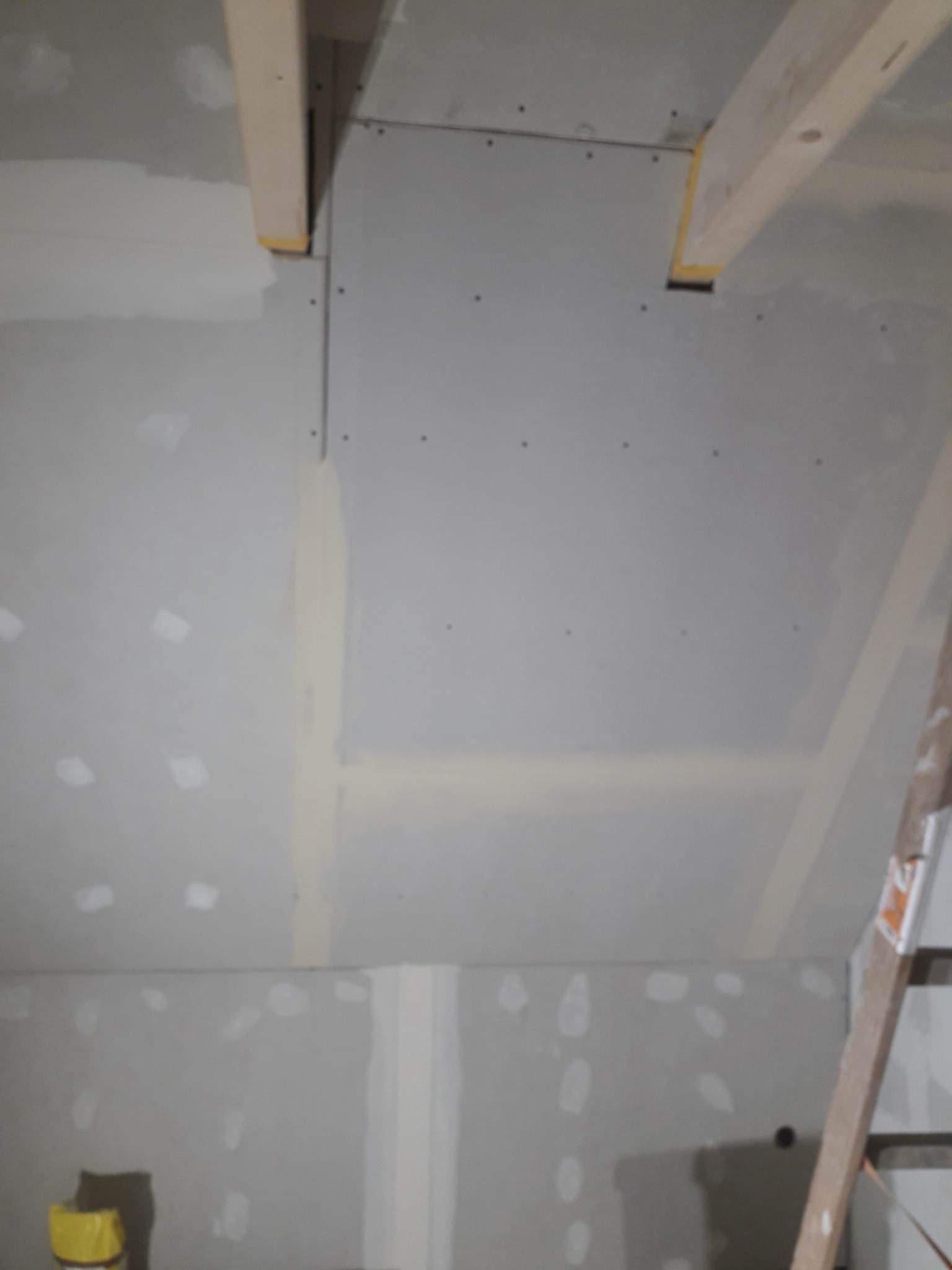 Skośna ściana poddasza z płyt gipsowo-kartonowych, widoczne drewniane belki stropowe z żółtą taśmą zabezpieczającą, szpachlowane łączenia płyt, drabina oparta o ścianę.