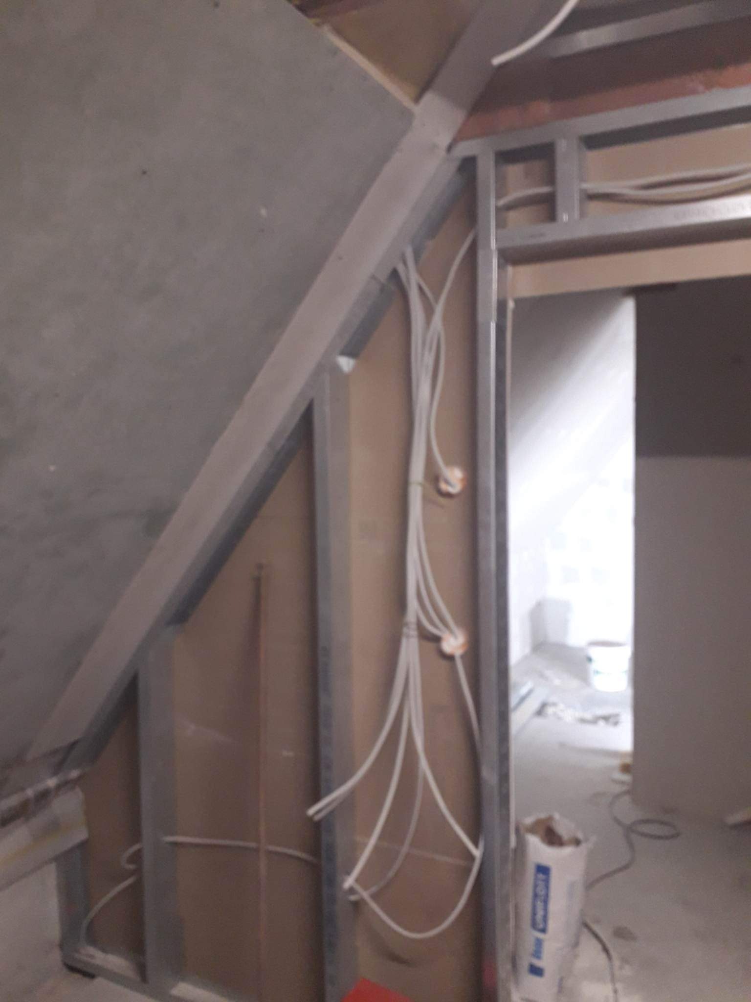 Szkielet ścianki działowej z profili metalowych w trakcie montażu, widoczne przewody elektryczne w peszlach i częściowo położona płyta gipsowo-kartonowa na poddaszu.