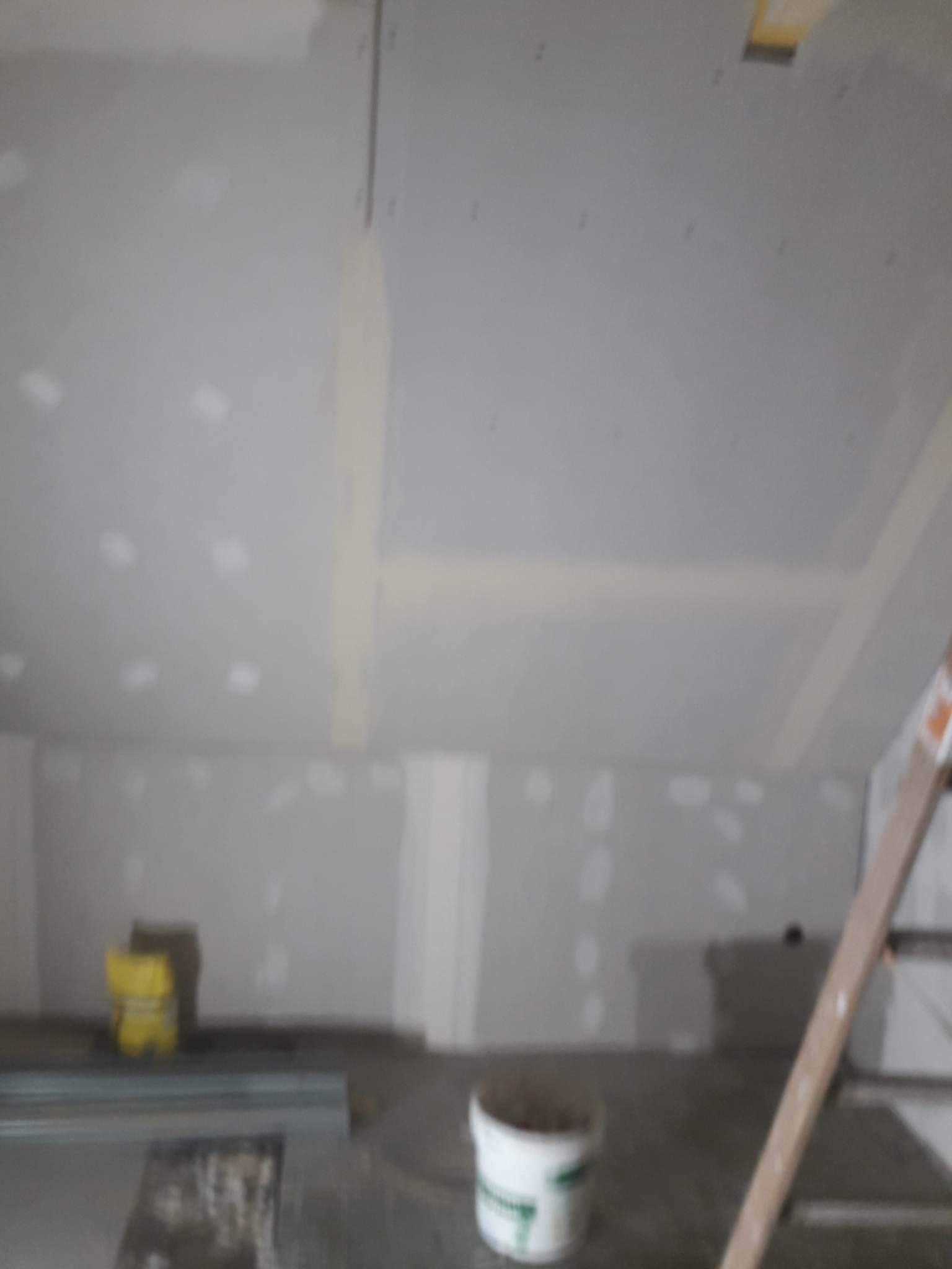 Wnętrze w trakcie prac wykończeniowych, widoczne płyty gipsowo-kartonowe na ścianach i suficie, częściowo zaszpachlowane łączenia, na podłodze wiadro i drabina.