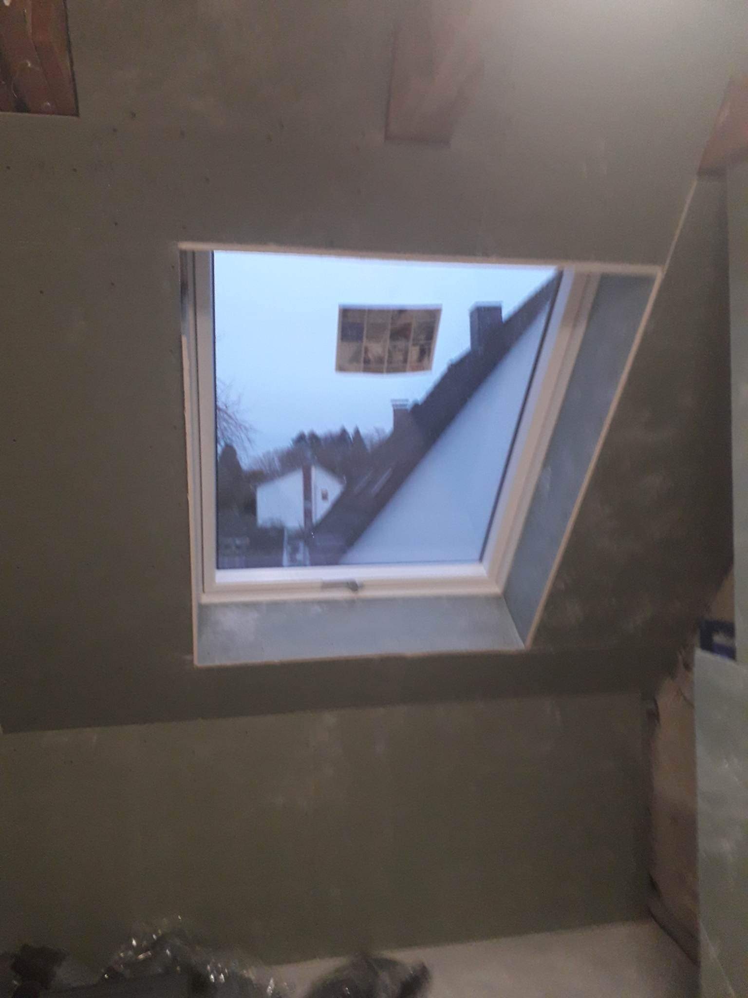 Montaż okna dachowego w surowym wnętrzu poddasza z widokiem na domy i drzewa na zewnątrz.