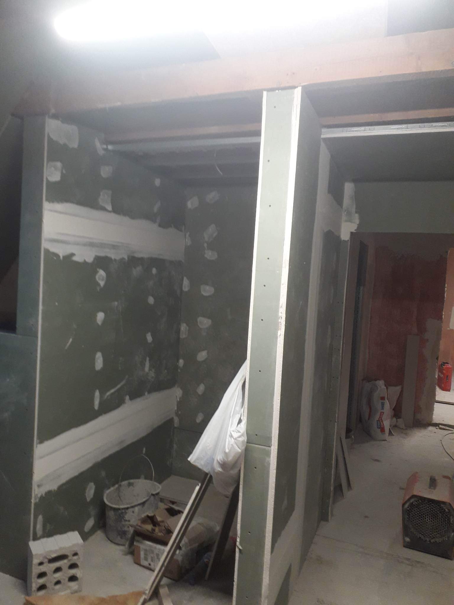 Wnętrze w trakcie prac wykończeniowych: surowe ściany z płyt gipsowo-kartonowych, widoczne łączenia szpachlowane, sufit z drewnianymi belkami, prowizoryczne oświetlenie, wiadro i narzędzia...