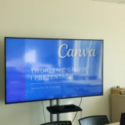 Ekran telewizora prezentujący tytuł szkolenia 'Canva - tworzenie grafik i prezentacji' prowadzonego przez Gabrielę Woch w dniach 16-17.03.2023. W tle widoczny fragment sali konferencyjnej...