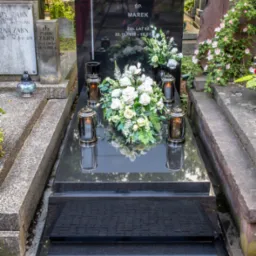 Czarny, polerowany nagrobek z inskrypcją 'ŚP. MAREK ŻYŁ LAT 62 22.11.1958-22.01', ozdobiony białymi kwiatami, zniczami i krzyżem, na tle cmentarza.