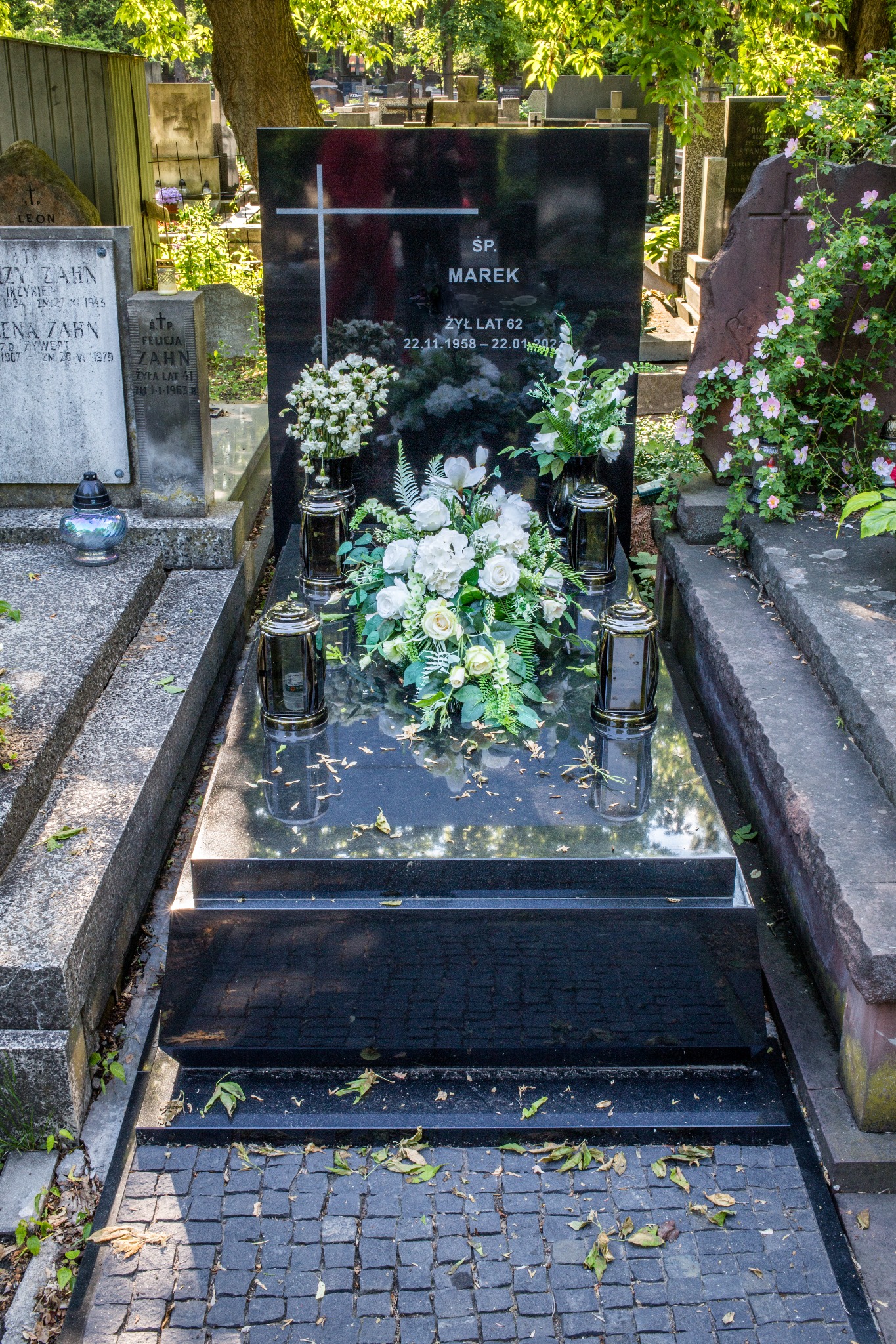 Czarny, polerowany pomnik z białymi kwiatami i lampionami, otoczony innymi nagrobkami na cmentarzu, z widocznymi liśćmi na ziemi.