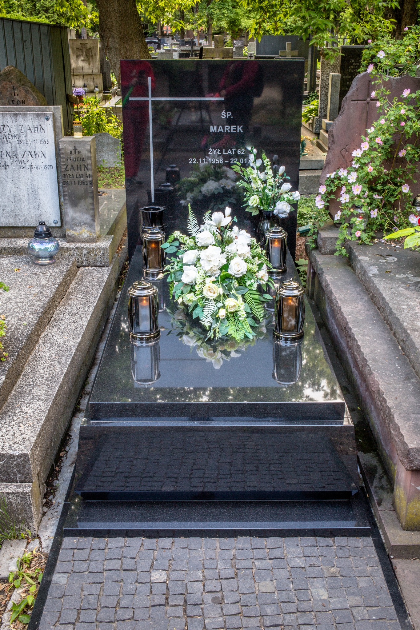 Czarny, polerowany nagrobek z inskrypcją 'ŚP. MAREK ŻYŁ LAT 62 22.11.1958-22.01', ozdobiony białymi kwiatami, zniczami i krzyżem, na tle cmentarza.