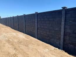 Długi, ciemnoszary mur z betonowych bloczków z jasnymi, betonowymi słupkami, biegnący wzdłuż piaszczystego terenu pod błękitnym niebem.