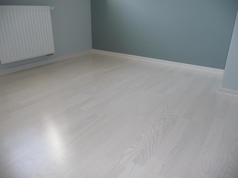 Jasne, bielone panele podłogowe z widocznym rysunkiem drewna, ułożone w pustym pokoju z błękitnymi ścianami i białym grzejnikiem.