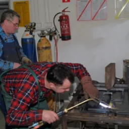 Pracownik w czerwonej koszuli w kratę spawa metalową konstrukcję palnikiem acetylenowo-tlenowym, drugi pracownik w tle obserwuje, butle z gazem w tle, instrukcje BHP na ścianie.