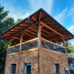 Dwukondygnacyjna konstrukcja z cegły i drewna z ażurową balustradą, zwieńczona drewnianym zadaszeniem na tle błękitnego nieba