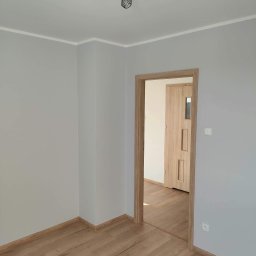 Michał K - Pomalowane na szaro wnętrze pokoju z drewnianą podłogą i ościeżnicą, widoczna lampa sufitowa z trzema reflektorami i drzwi do kolejnego pomieszczenia.