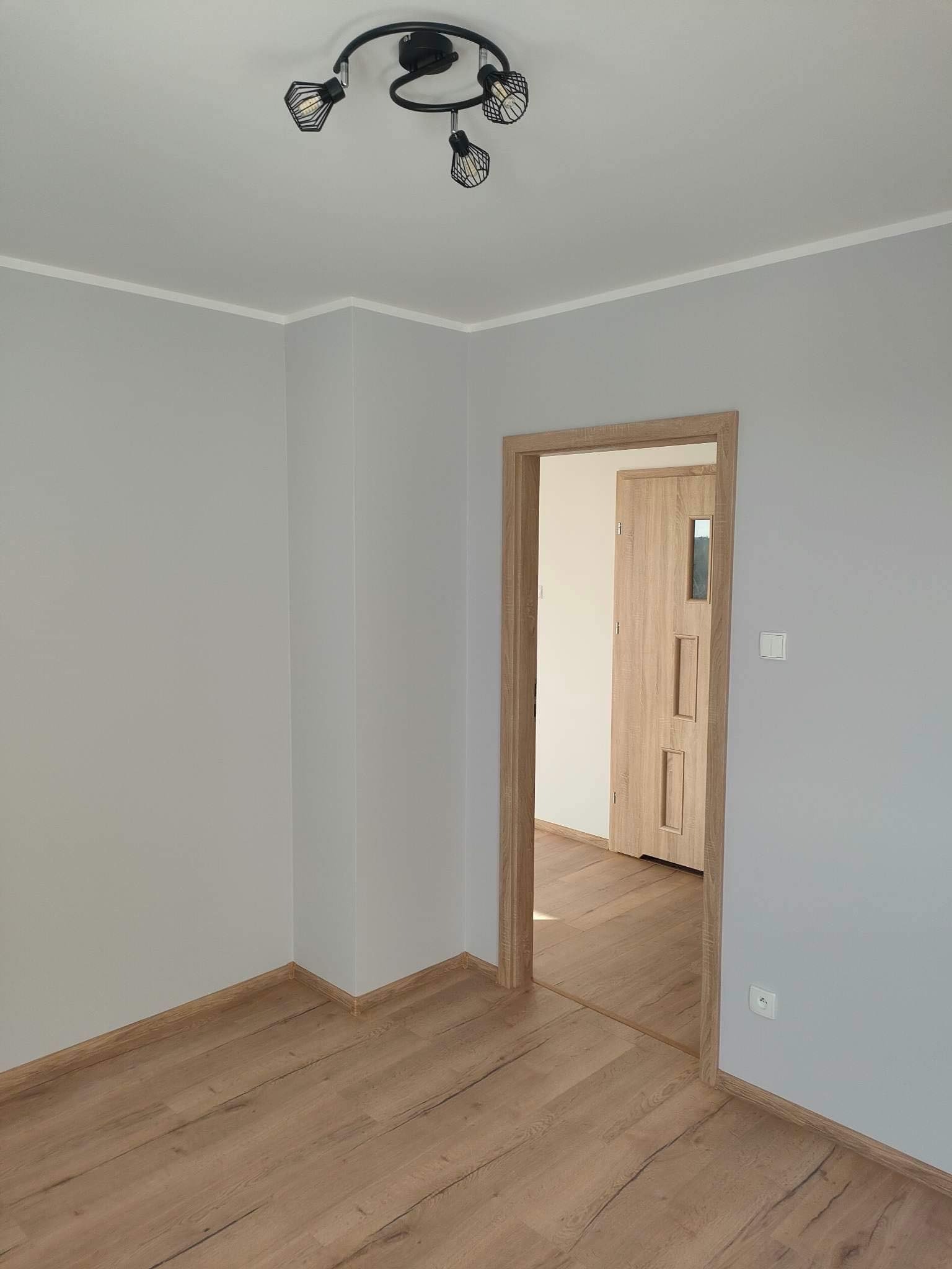 Pomalowane na szaro wnętrze pokoju z drewnianą podłogą i ościeżnicą, widoczna lampa sufitowa z trzema reflektorami i drzwi do kolejnego pomieszczenia.