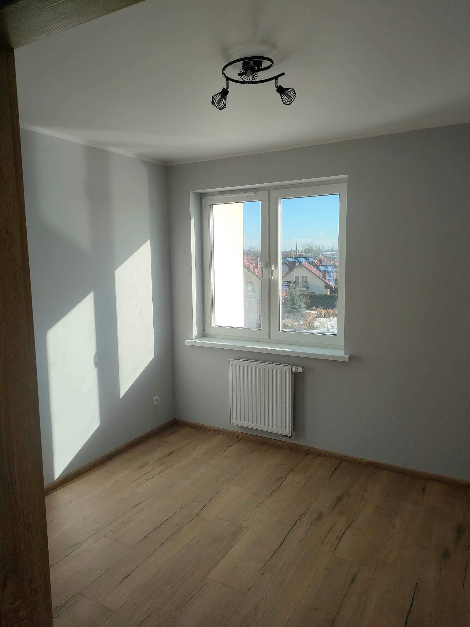 Pomalowane na szaro puste pomieszczenie z drewnianą podłogą, białym grzejnikiem i widokiem na domy za oknem.