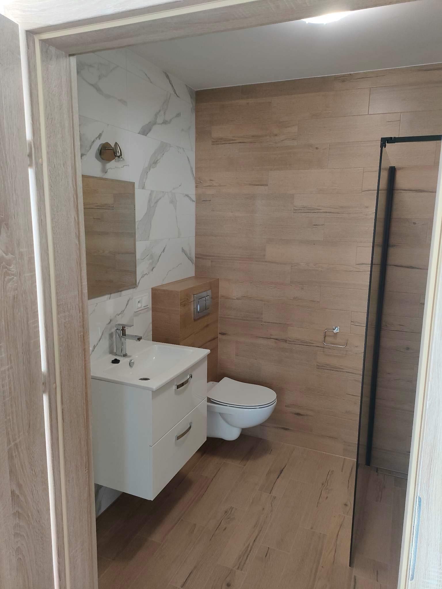 Nowoczesna łazienka z białymi płytkami imitującymi marmur, drewnopodobnymi płytkami na pozostałych ścianach i podłodze, białą szafką pod umywalką z szufladami, wiszącą toaletą i kabiną prysznicową...