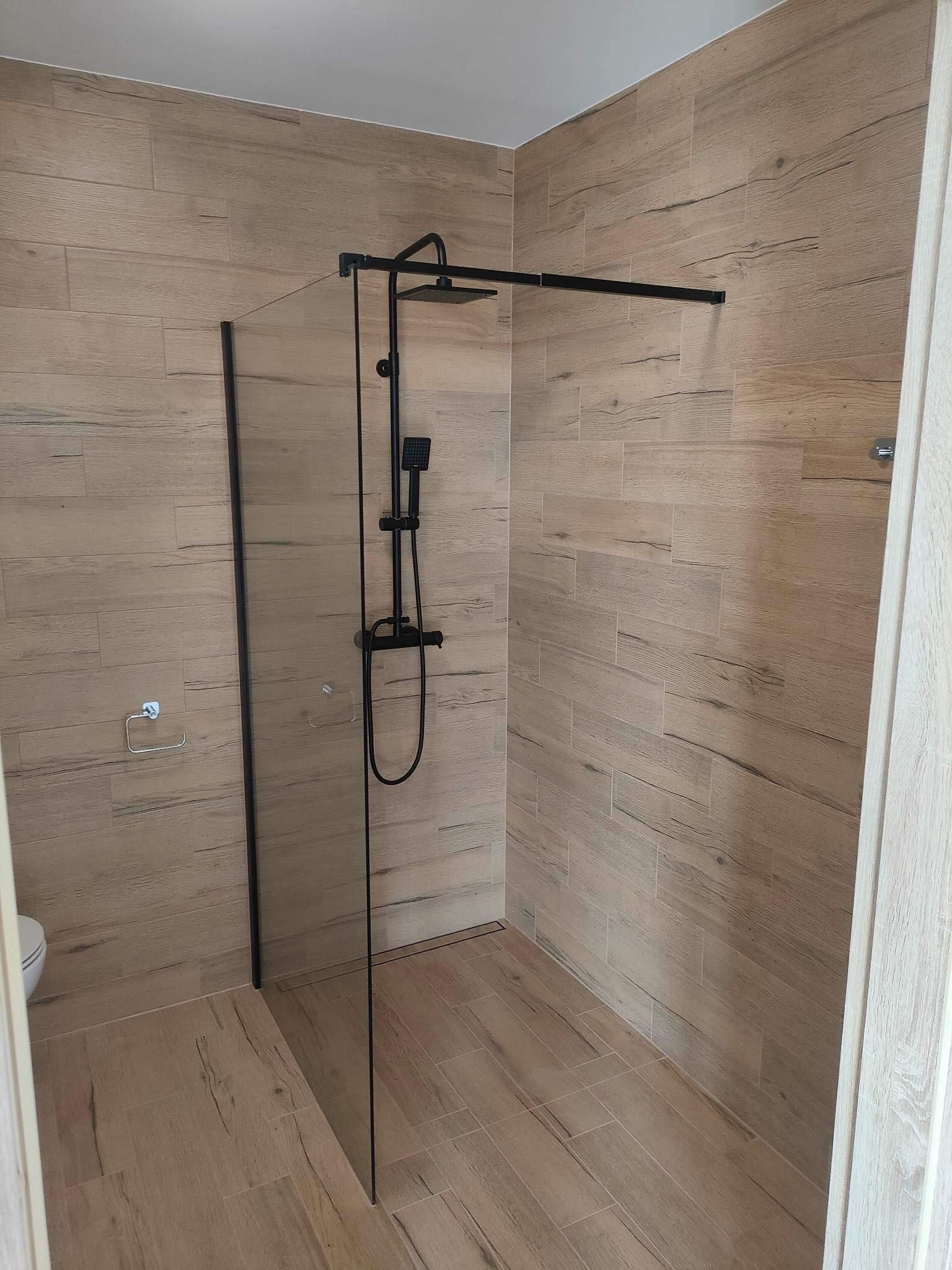 Nowoczesna kabina prysznicowa walk-in z czarną armaturą i okładziną ścienną imitującą drewno, z widocznym odpływem liniowym w podłodze.