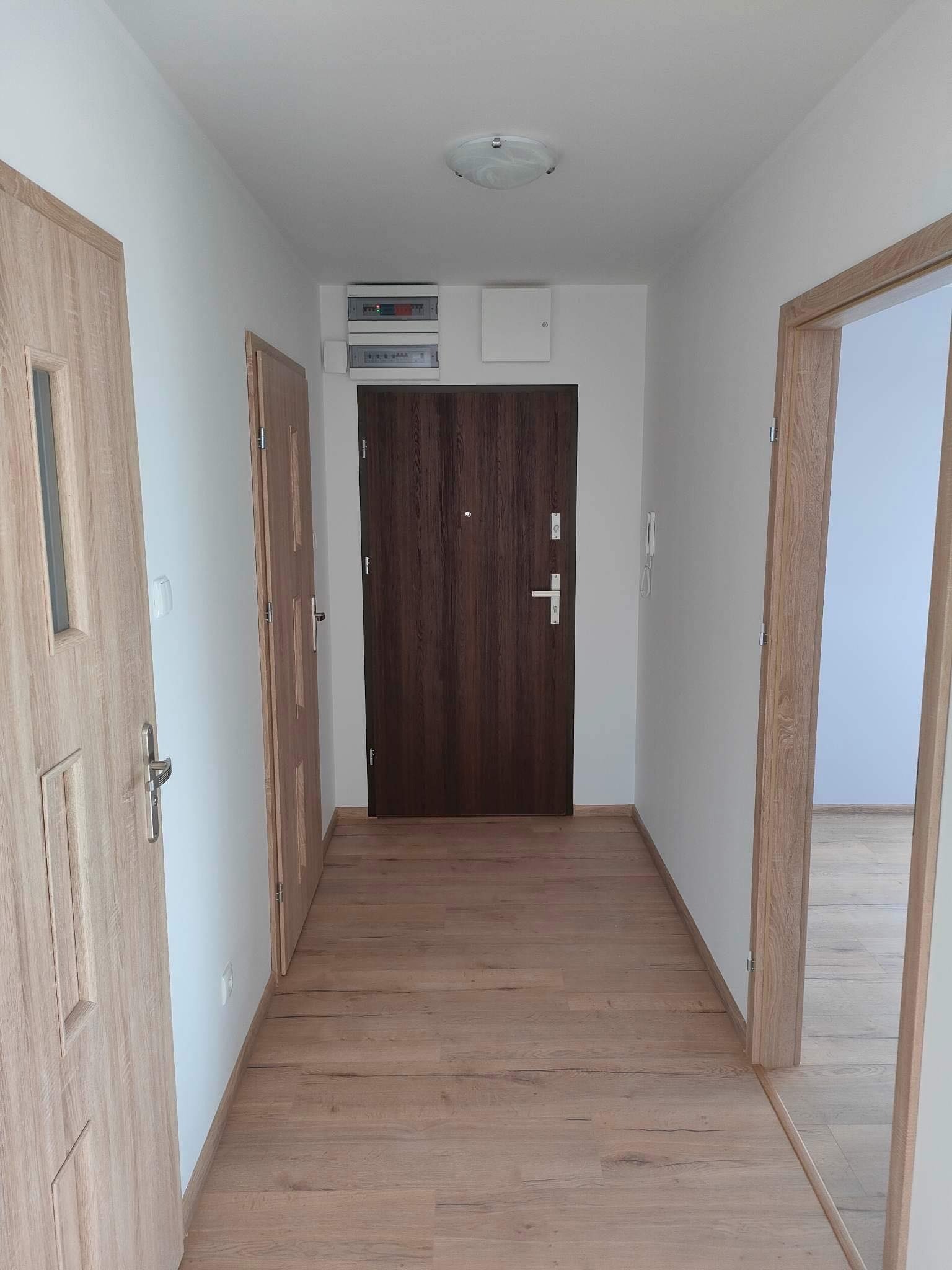 Korytarz po remoncie z drewnianą podłogą, dwiema parami drzwi z tego samego materiału i brązowymi drzwiami wejściowymi.