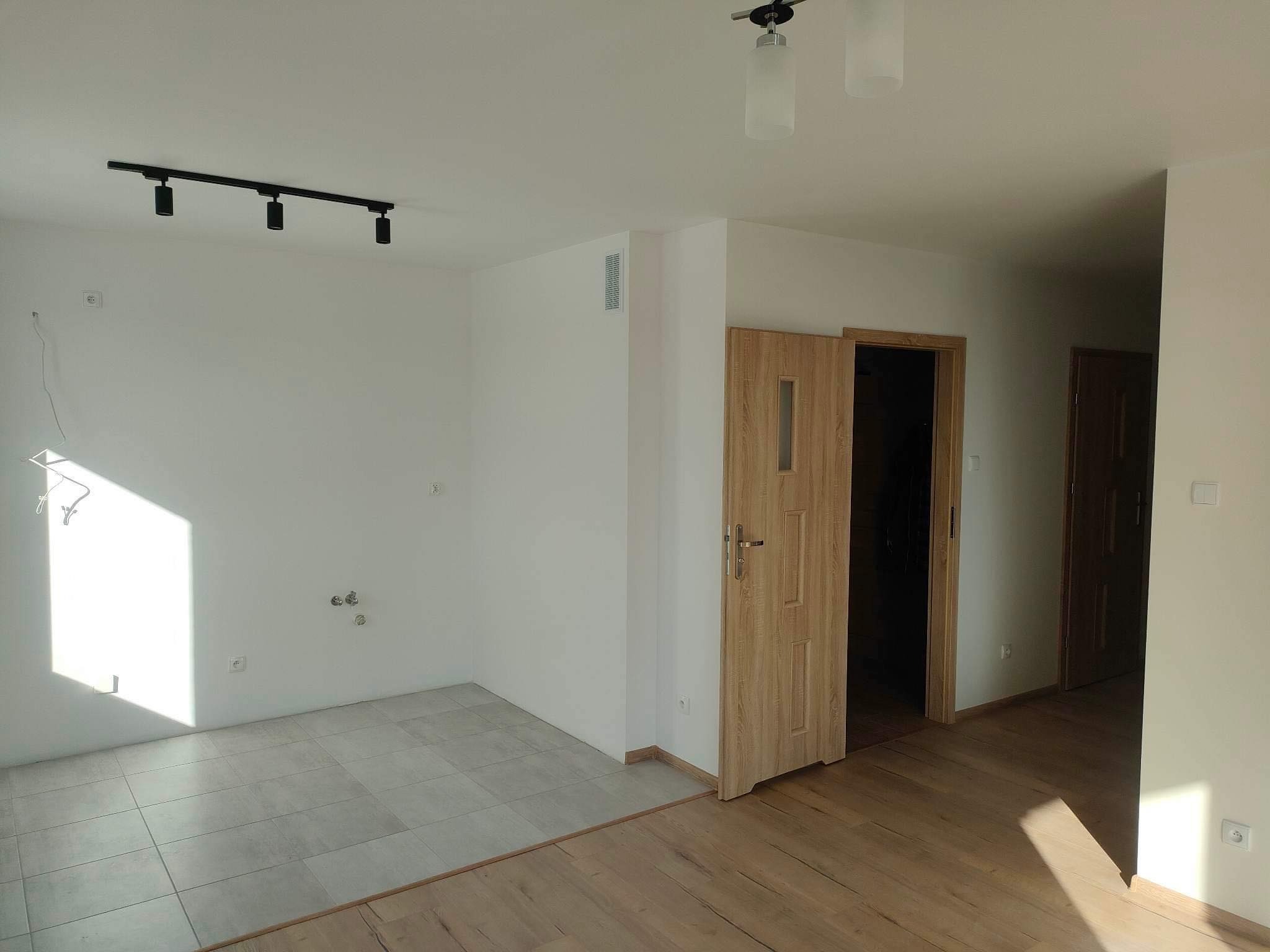 Wnętrze pokoju po remoncie z dwiema różnymi podłogami: płytki ceramiczne i panele drewniane, białe ściany z czarną lampą sufitową i drewnianymi drzwiami.