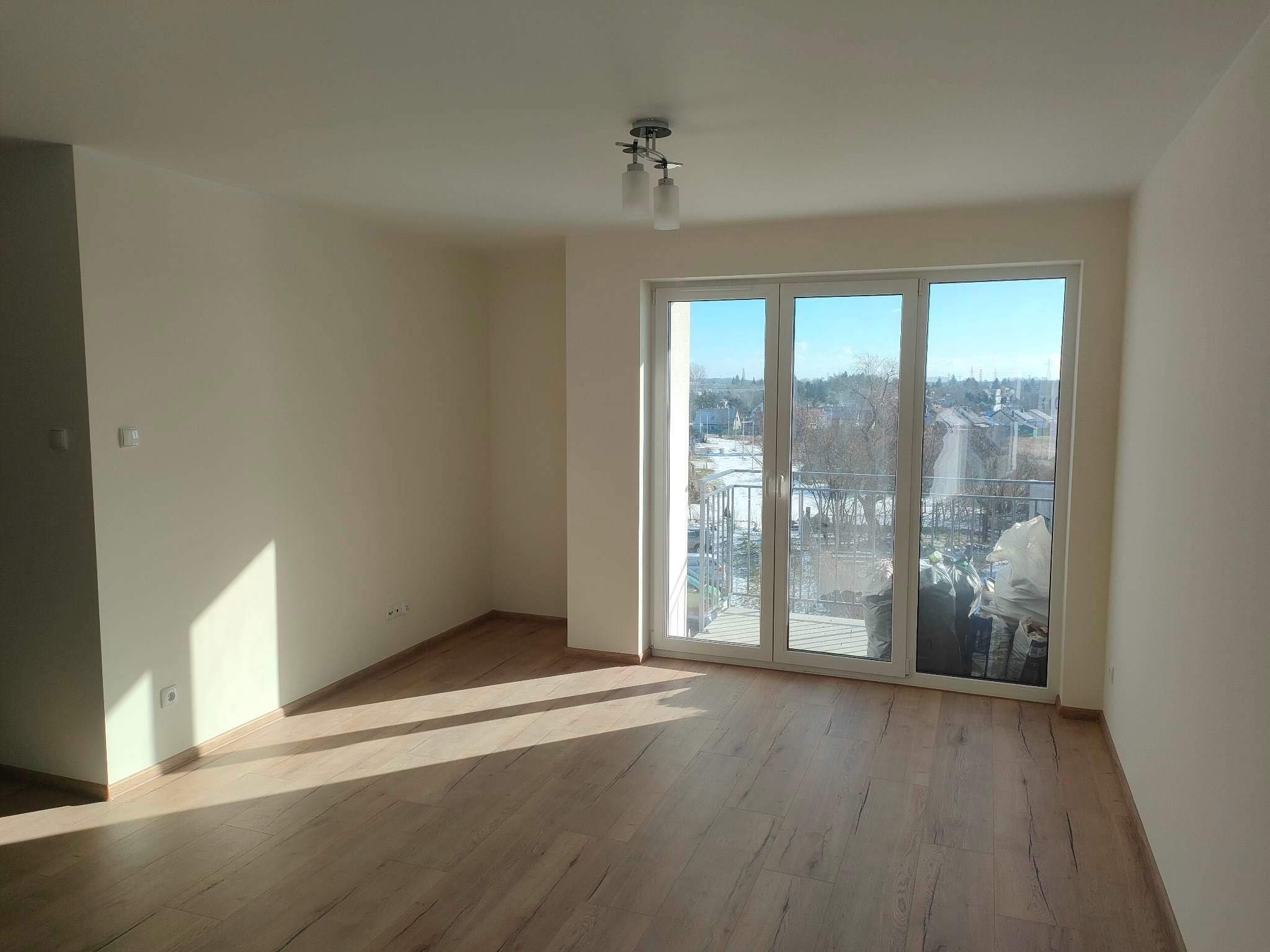 Jasne, puste pomieszczenie z drewnianą podłogą i balkonem z widokiem na miasto w słoneczny dzień. Świeżo pomalowane ściany w odcieniach beżu.