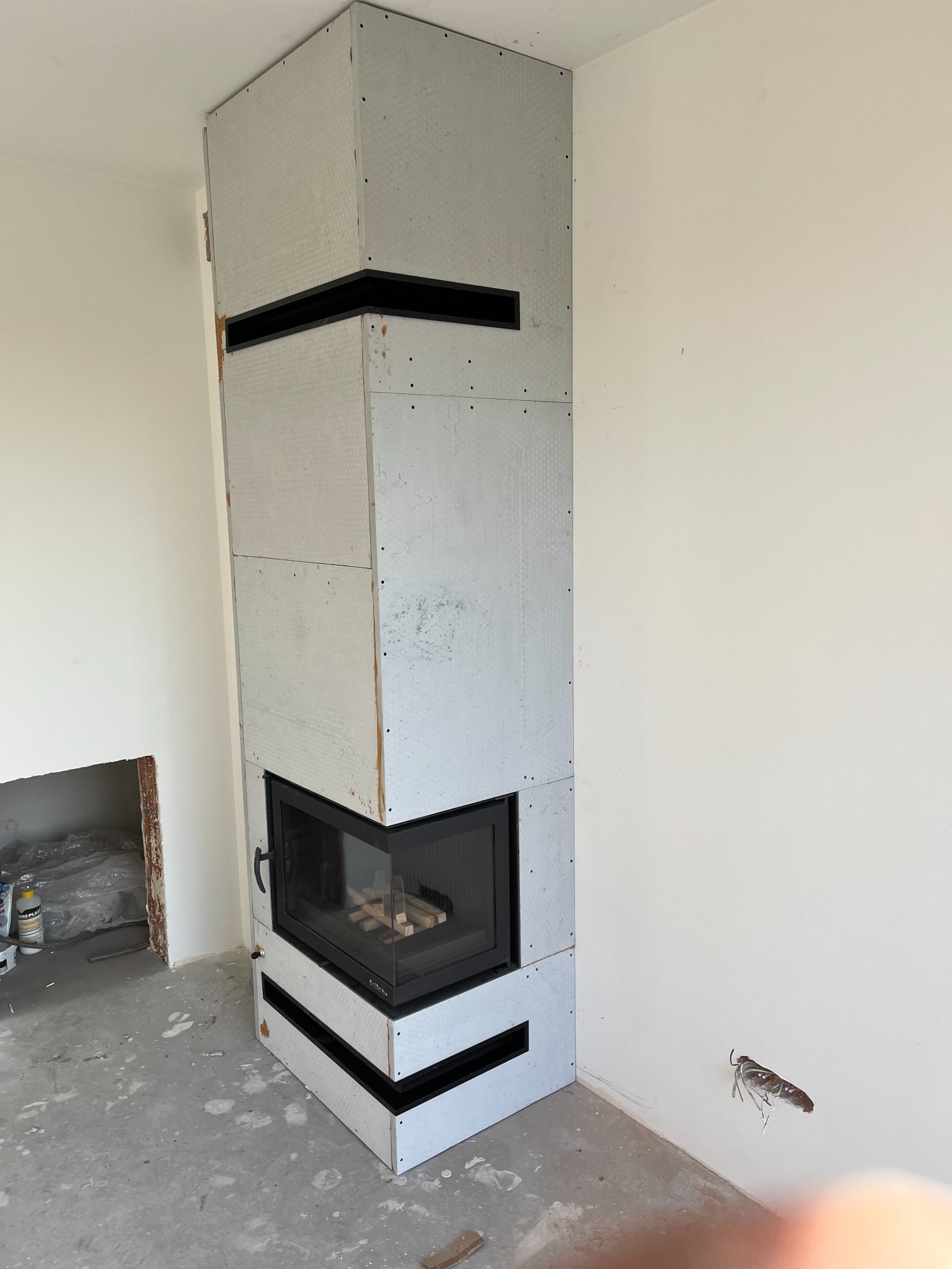 Wnętrze w trakcie montażu obudowy wkładu kominkowego z płyt gipsowo-kartonowych z widocznymi czarnymi elementami dekoracyjnymi, surowy betonowy podkład podłogowy.