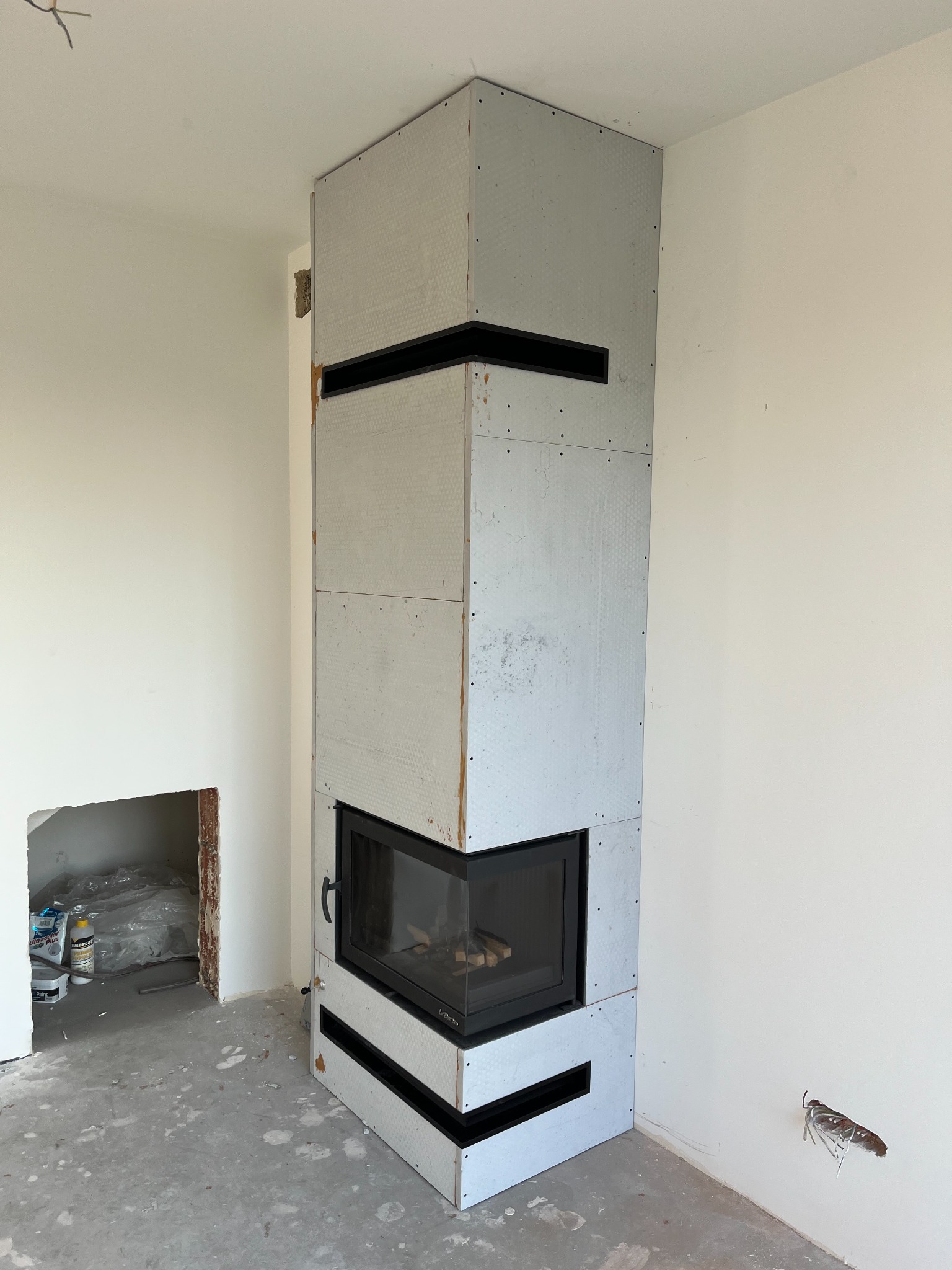 Wnętrze w trakcie wykończenia z zabudową wkładu kominkowego z płyt karton-gips, widoczne czarne wloty powietrza i otwór na wkład kominkowy w ścianie obok.
