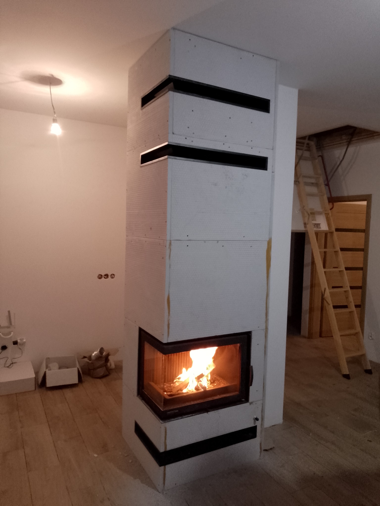 Wnętrze z nowoczesnym narożnym wkładem kominkowym obudowanym płytami gipsowo-kartonowymi z czarnymi akcentami wentylacyjnymi, w tle drewniana drabina oparta o ścianę.