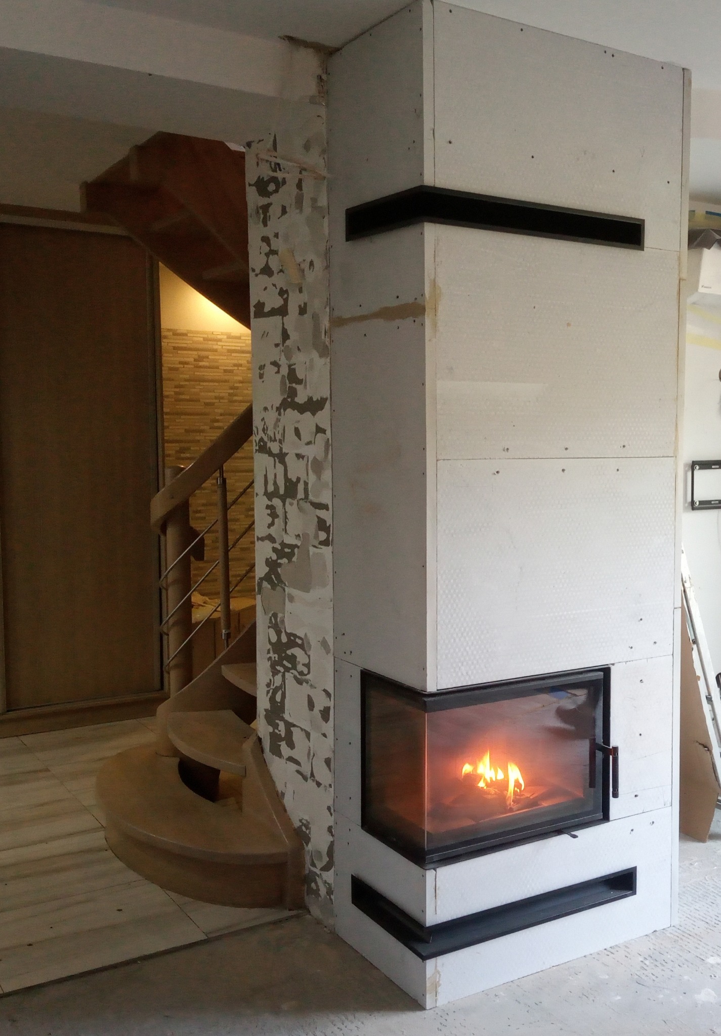 Wnętrze w trakcie wykończenia: obudowa wkładu kominkowego z płyt gipsowych z czarnymi elementami dekoracyjnymi, widoczne płomienie, obok kręte schody drewniane i ściana z resztkami farby.