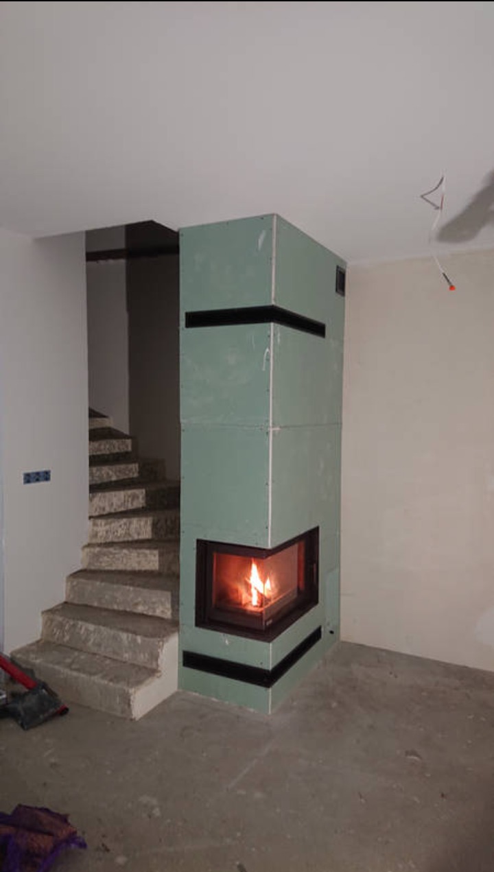 Niedokończona obudowa narożnego wkładu kominkowego z płyt gipsowo-kartonowych, widoczne czarne taśmy wzmacniające, obok surowe betonowe schody, wnętrze w trakcie remontu.